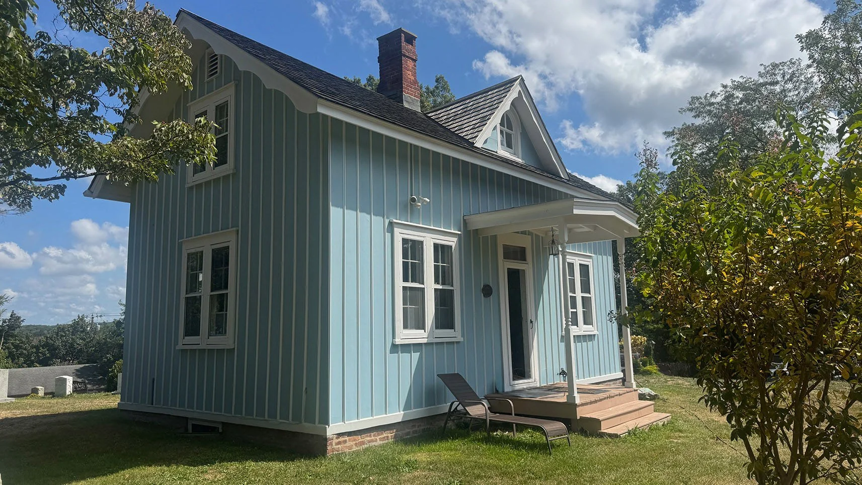A two-story light blue house with white trim and a brick foundation, featuring a front porch, multiple windows, a brick chimney, surrounded by green grass and trees on a sunny day with some clouds in the sky.