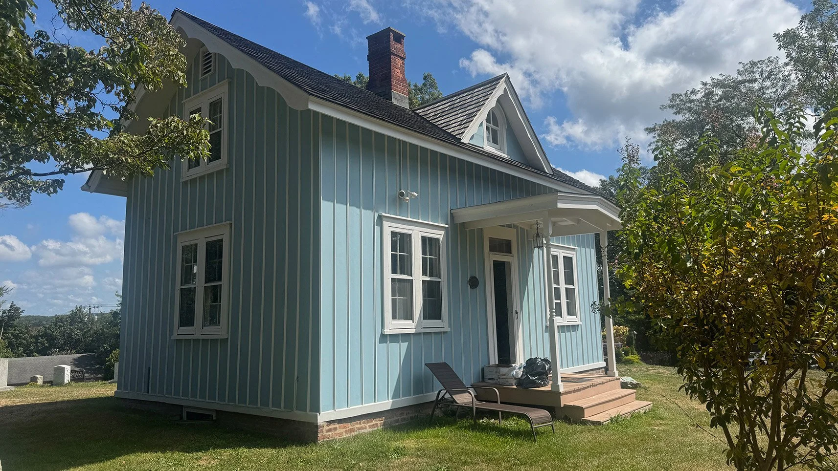 A light blue, two-story house with vertical white stripes, a gabled roof, and a small front porch with stairs, set in a grassy yard with trees and a partly cloudy sky.
