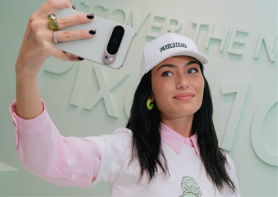 Young woman with dark hair, wearing a white cap and pink and white shirt, holding a smartphone while taking a selfie