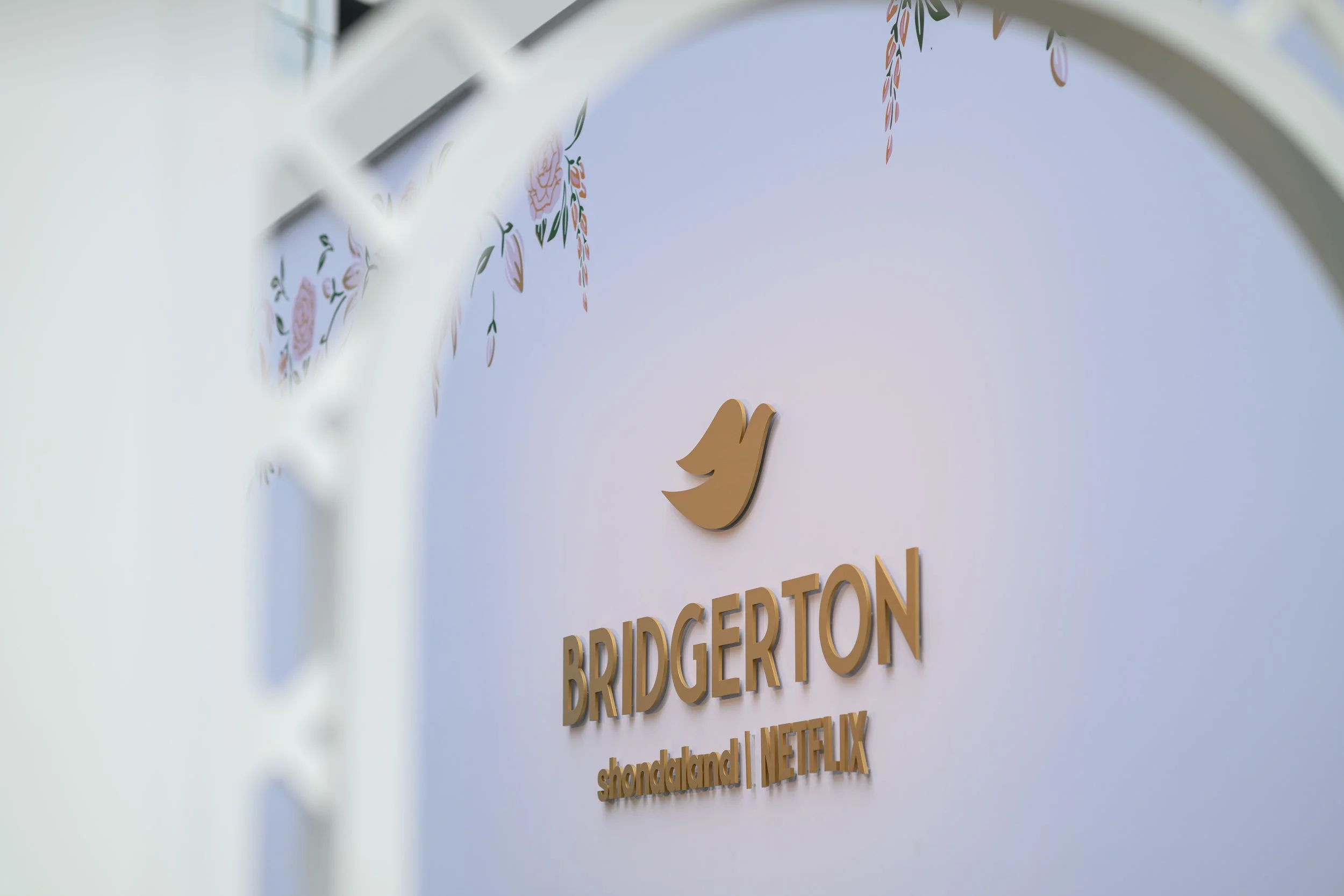 A sign with the Twitter bird logo and text that reads 'BRIDGERTON shondaland | NETFLIX' on a wall, partially framed by a blurred white lattice in the foreground, and with a floral mural above.