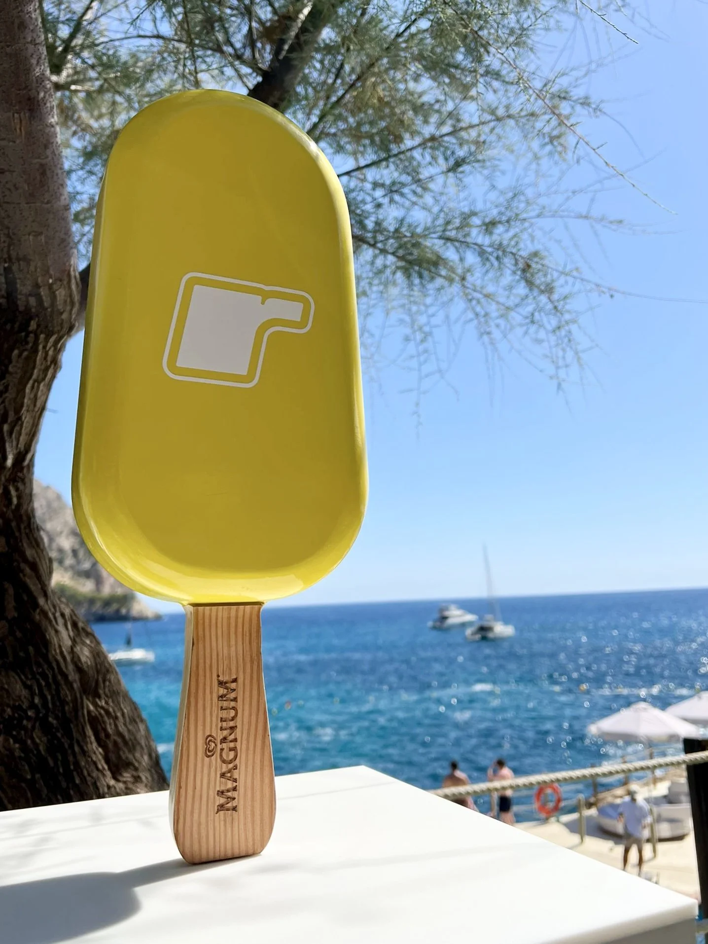 A yellow and wooden magnum ice cream bar, with a white outline of a popsicle on it, sits on a table near a beach with boats and people in the background, under a tree and clear blue sky.