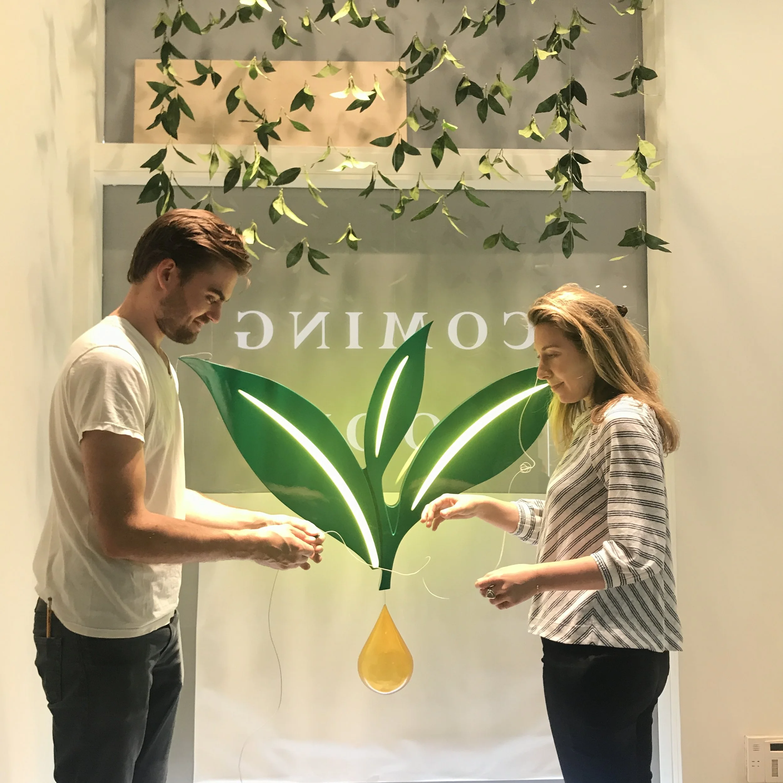 Two people, a man and a woman, standing in front of a wall with a large illuminated leaf design with a drop shape at the bottom. They are holding wires and appear to be working on the installation.