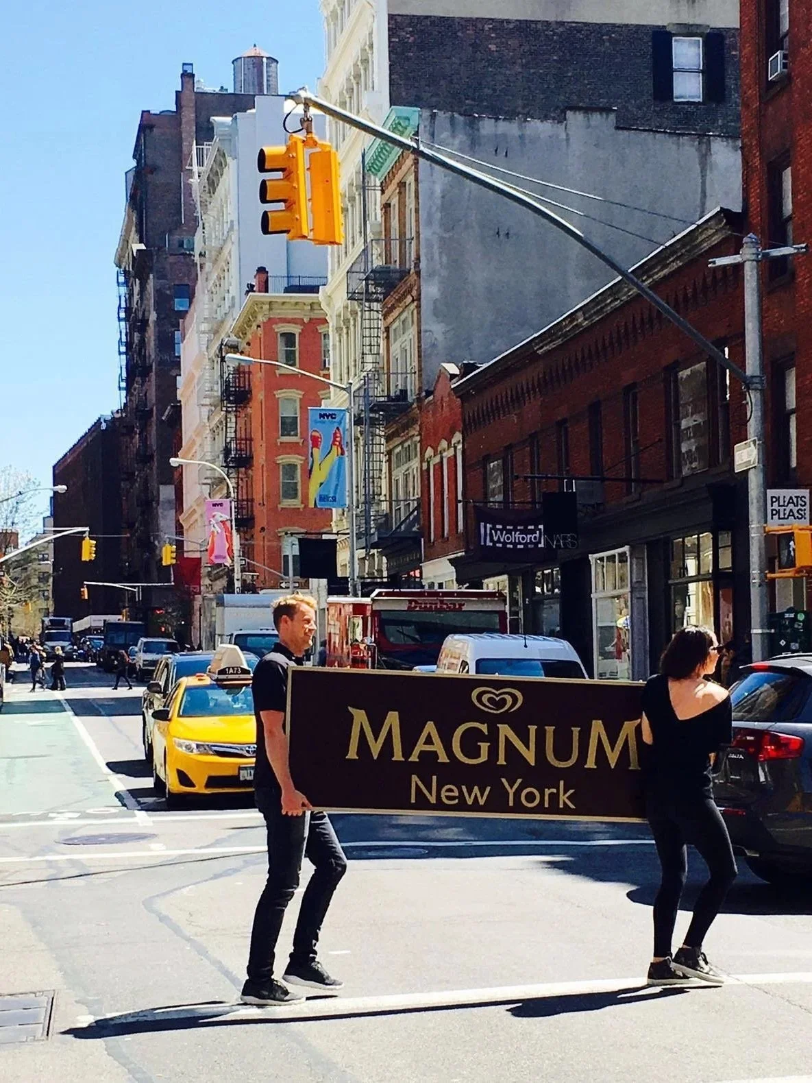 Two people walking across a city street, holding a large sign that reads 'MAGNUM New York'. Cars and buildings line the street, with a traffic light overhead and a clear blue sky.
