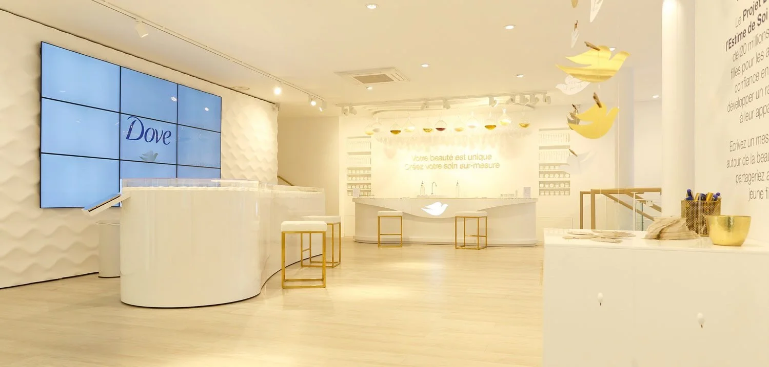 A bright, modern perfume or cosmetics store with beige walls and flooring, featuring a Dove advertisement on a large digital display, a curved white counter, and white and gold decor. There are hanging yellow and white bird cutouts and shelves with products along the walls.