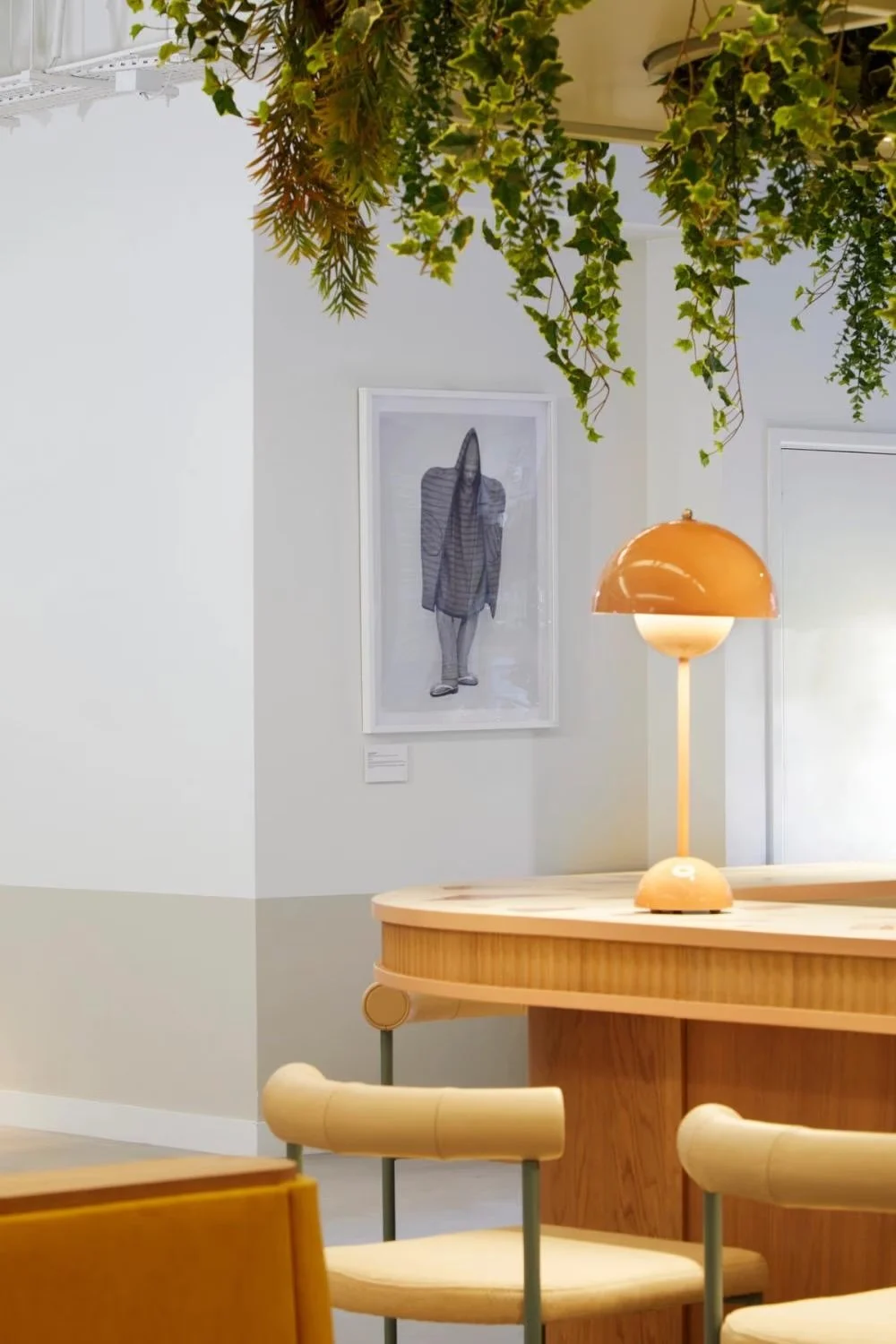 Interior of a modern, minimalist room with a wooden table, beige chairs, a sleek orange and white table lamp, and a framed black-and-white photo of a person on the white wall. Green leafy plants hanging from the ceiling add freshness to the space.