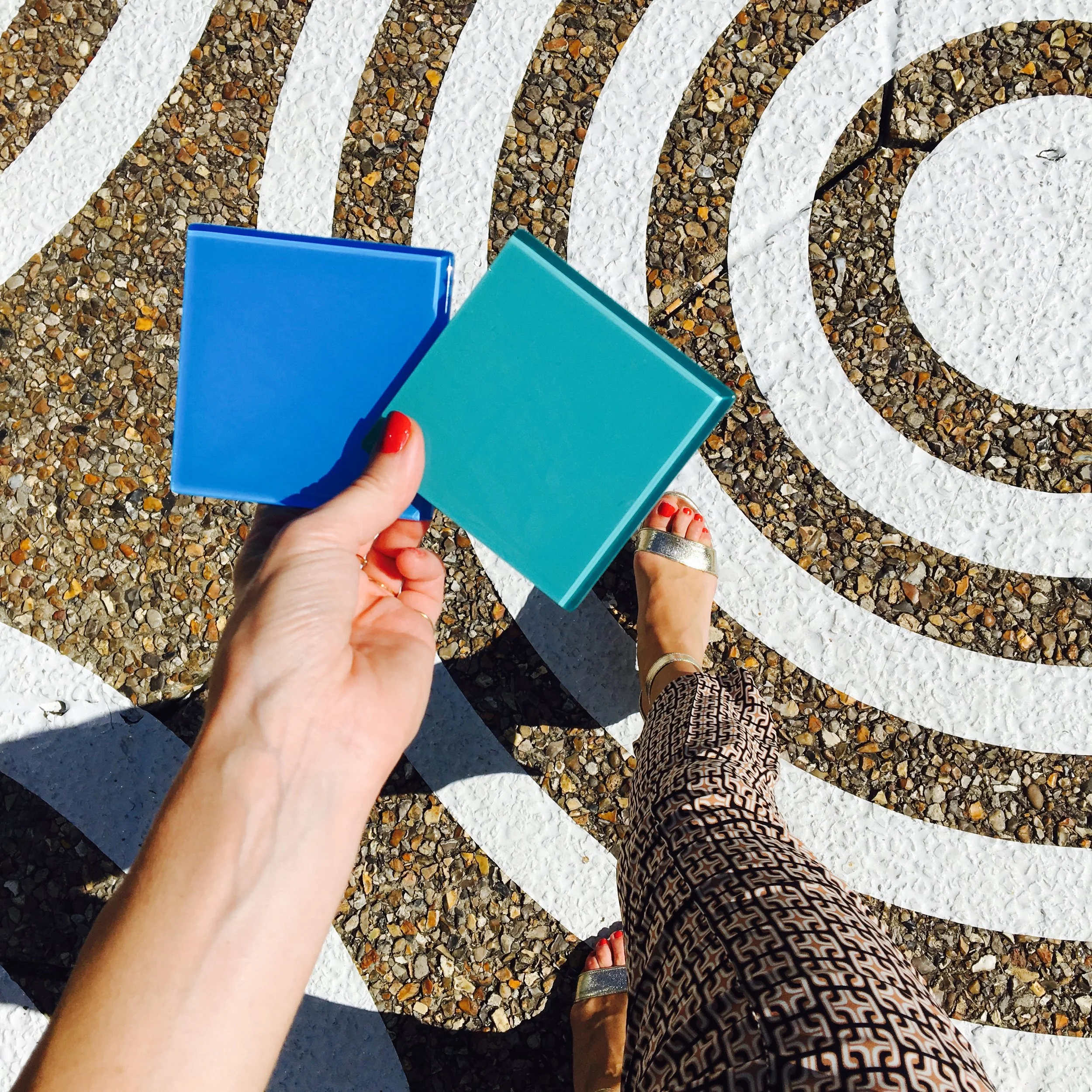 A person holding two square colored tiles, blue and teal, while standing on a textured surface with white circular patterns.