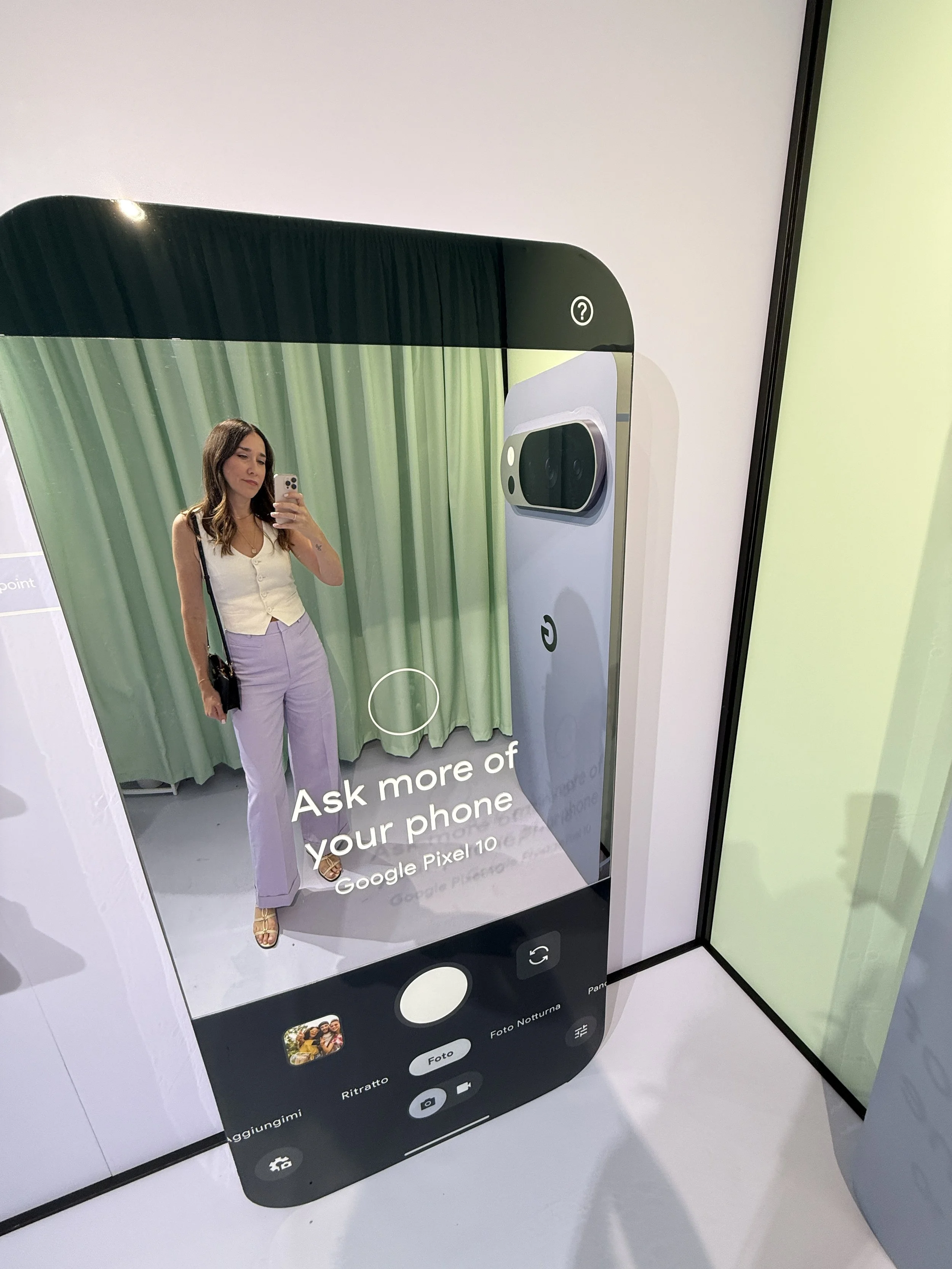 A woman in a white sleeveless top and lavender pants taking a mirror selfie in a cell phone display center. The display shows a large phone interface background with a green curtain and a smartphone graphic.