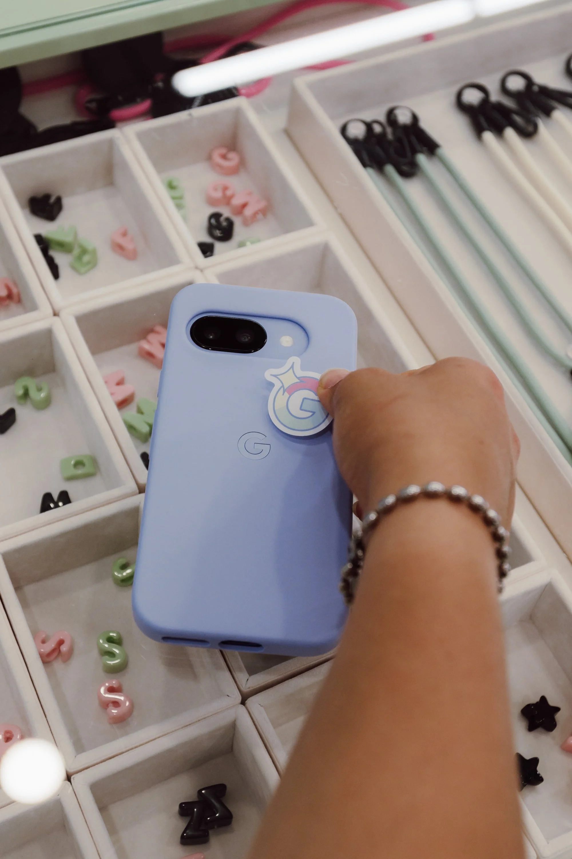 A person's hand holding a light blue Google Pixel phone with a sticker, over a display of colorful letter-shaped candies and black star-shaped candies in small white boxes, with striped straws in the background.