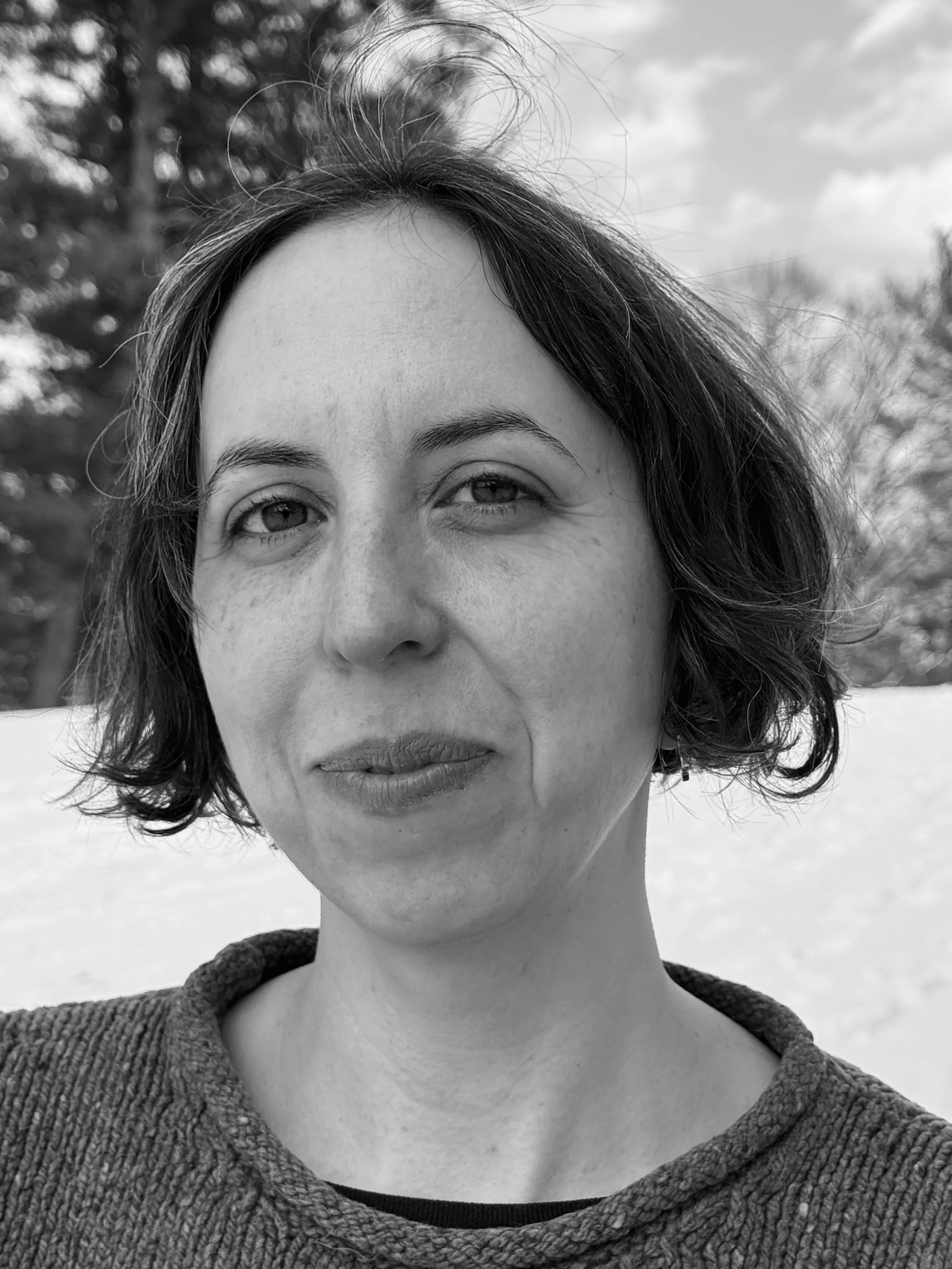 A black and white photo of a woman with short, wavy hair standing outdoors with trees in the background.