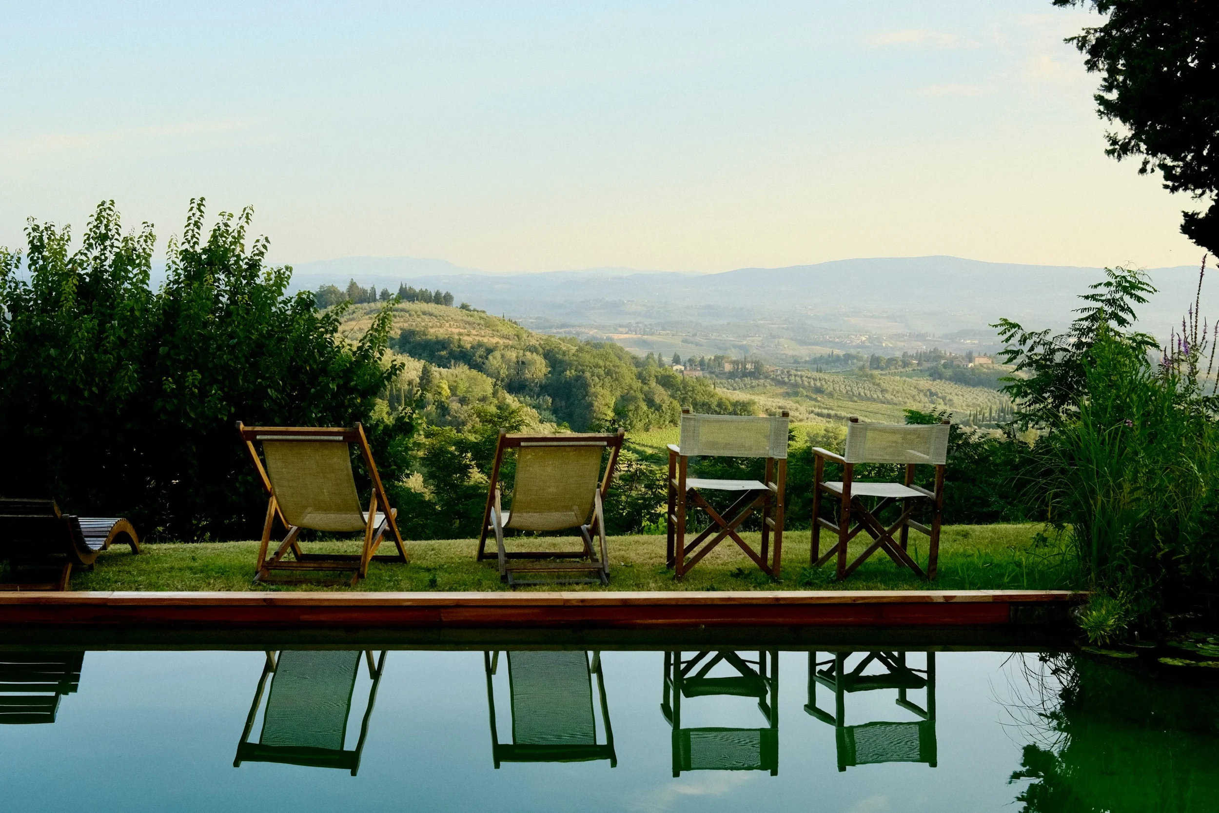 Scenic view of a row of six chairs and loungers facing a lush green landscape with rolling hills and distant mountains, reflected in a calm swimming pool.