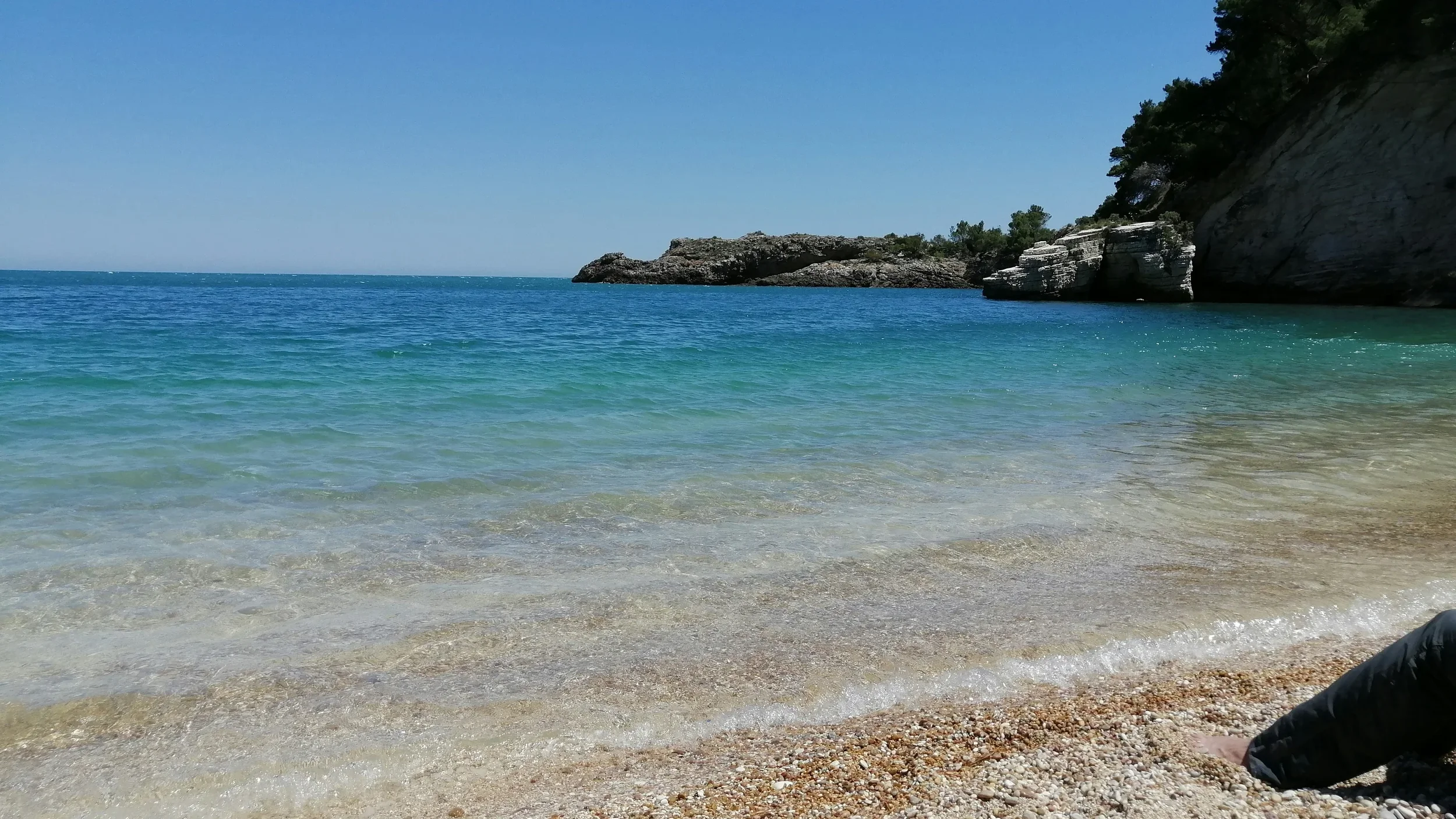 A serene beach scene with clear blue water lapping onto a pebbled shore, rocky cliffs in the background, and a bright blue sky overhead.