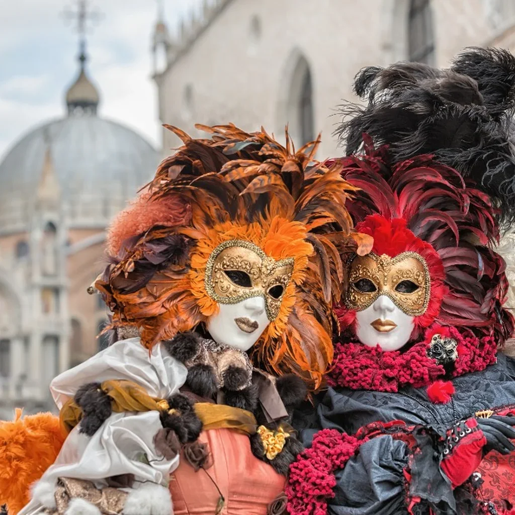 Two individuals in elaborate Venetian masks and costumes adorned with colorful feathers, standing outdoors in front of historical buildings during a festival or carnival.