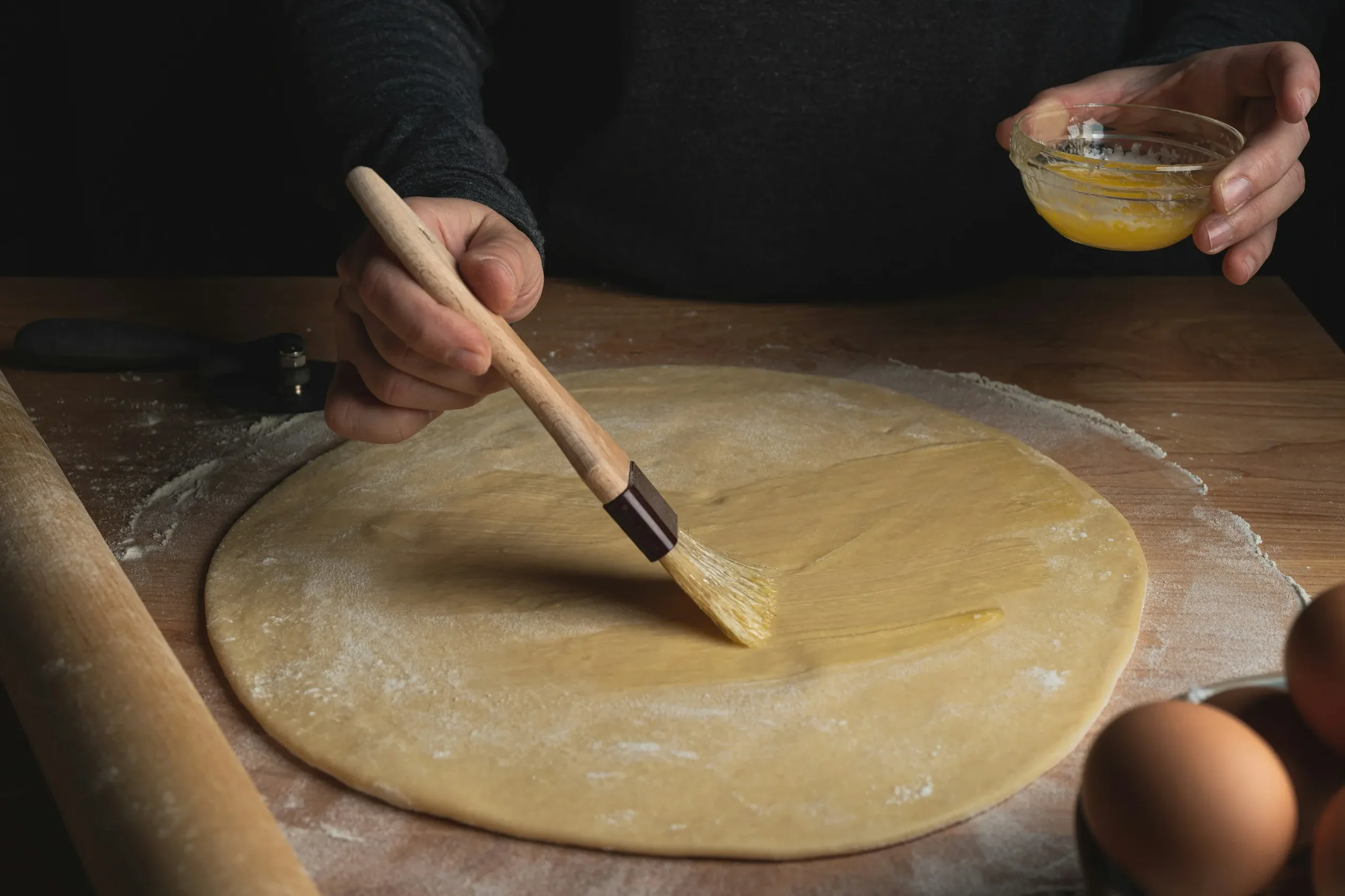 Person brushing rolled-out cookie dough on a wooden surface, with eggs and a rolling pin nearby, and holding a small bowl of egg wash.