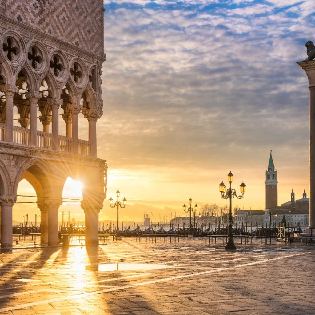 Sunset over a historic European cityscape with ornate architecture, lampposts, and a bell tower in the background, near a waterfront with boats.