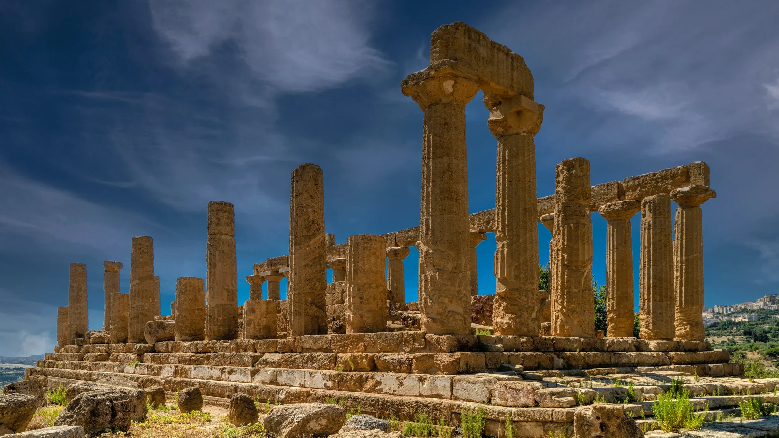 Ancient stone temple ruins with columns under a partly cloudy sky, located on a hillside.