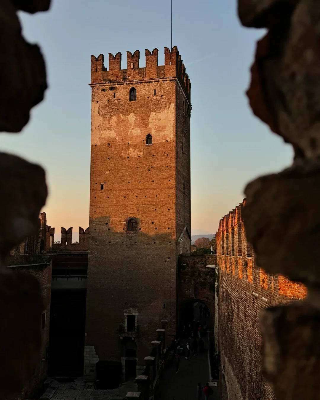 A tall medieval brick tower viewed through a small opening in a stone wall during sunset, with a clear sky in the background.