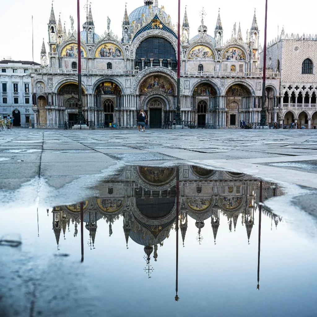 The image shows Saint Mark's Basilica in Venice, Italy, with its ornate domes and detailed facade. A puddle on the ground reflects the basilica's architecture, creating a mirror image.