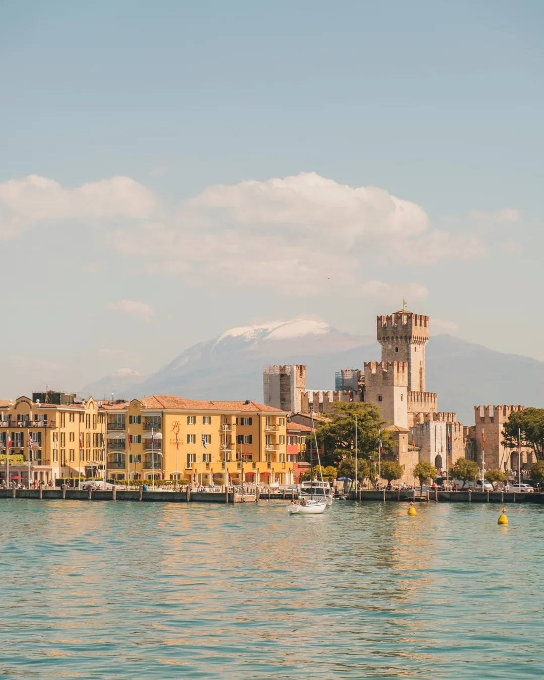 A marina with a sailboat near yellow buildings and a historic castle with a mountain in the background.
