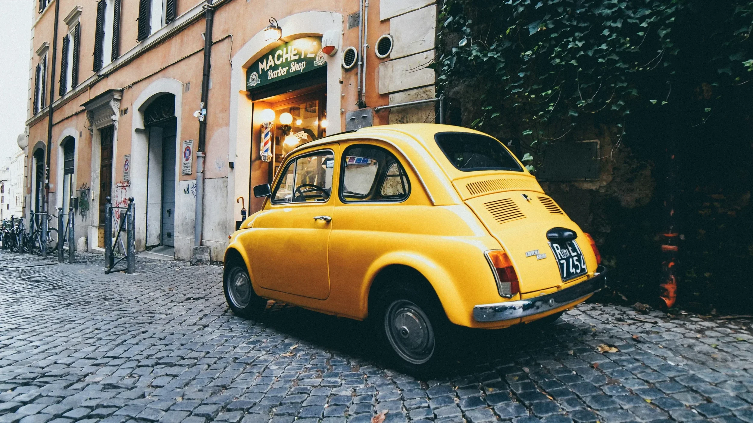 Yellow vintage Fiat 500 parked on cobblestone street near a barber shop with a sign reading 'MACHE Barber Shop' in a European city.