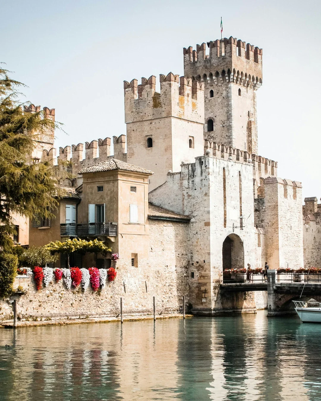 A historic castle with battlements and a tower, located on the water's edge, with reflection in the water and flowering plants nearby.