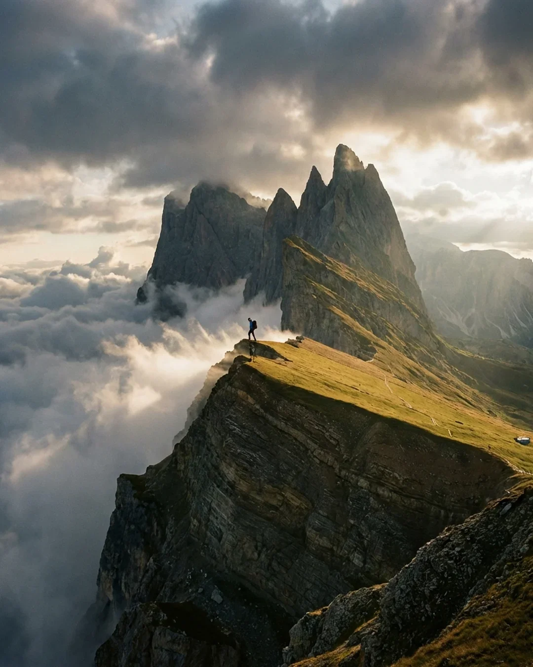 A hiker standing on a mountain ridge surrounded by clouds and fog, with towering rugged mountains in the background during sunrise or sunset.
