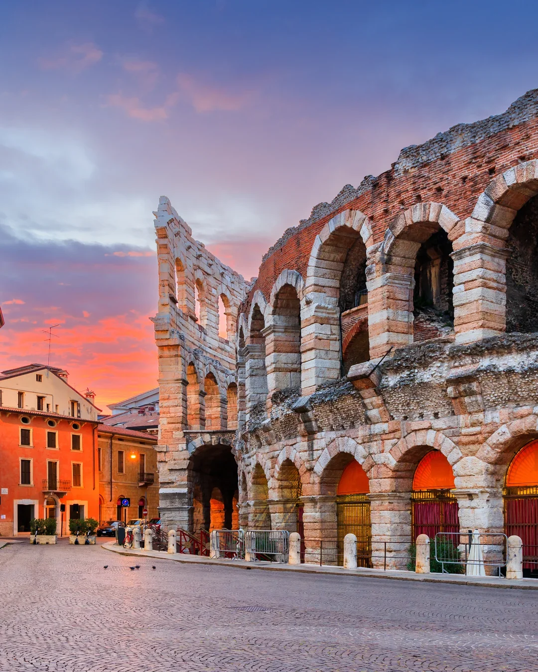 Ancient Roman amphitheater with brick arches during sunset, adjacent to colorful buildings in a cobblestone street.