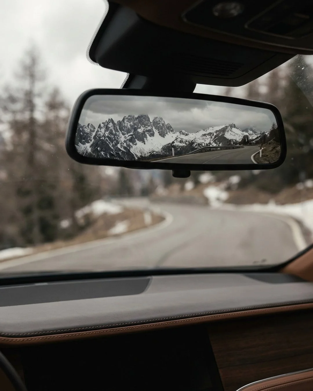 View from a car side mirror showing the reflection of snowy mountains and a winding road while leaving the Dolomites.
