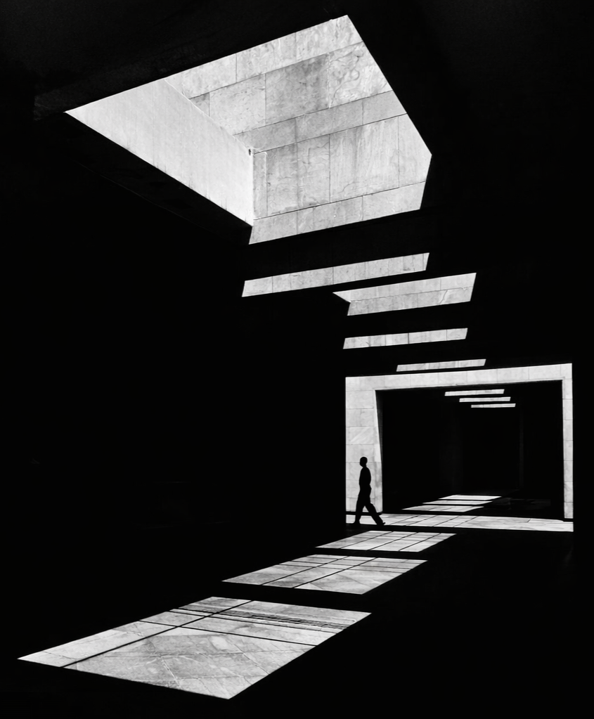A black and white photo of an indoor space with geometric shapes and strong contrast. Sunlight streams in through a large opening from above, creating patterns and shadows on the floor and walls. A small silhouette of a person walking inside.