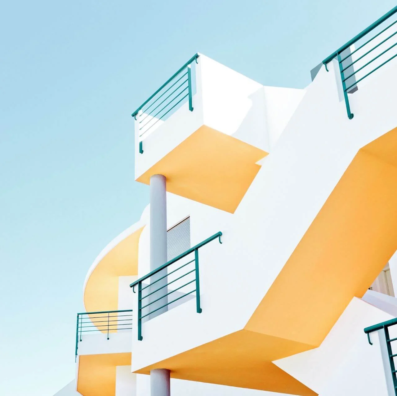 Modern white building with yellow accents and teal railings against a clear blue sky.