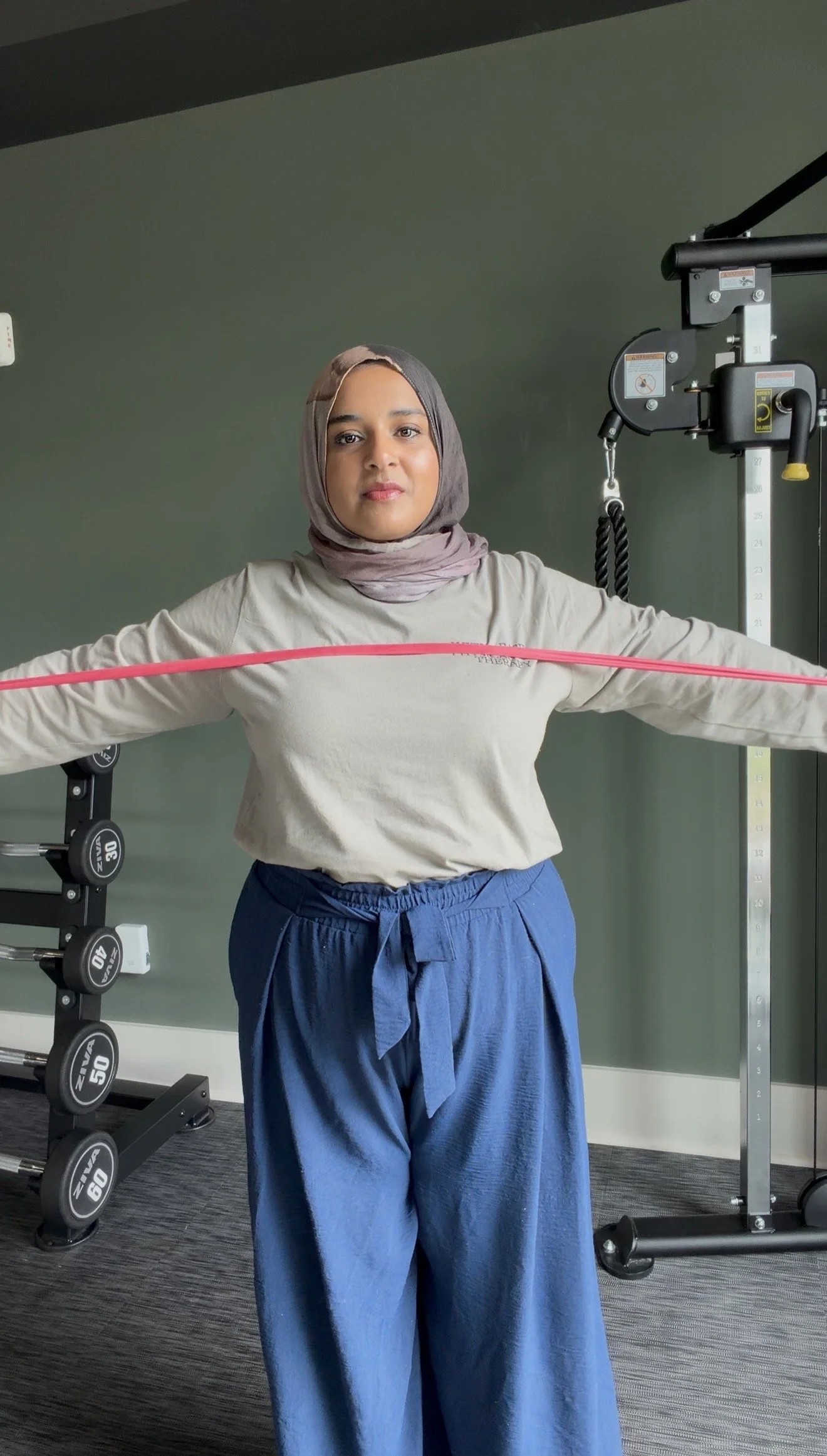 A woman wearing a hijab and loose-fit blue pants performs a stretching exercise with a pink resistance band across her chest in a gym.