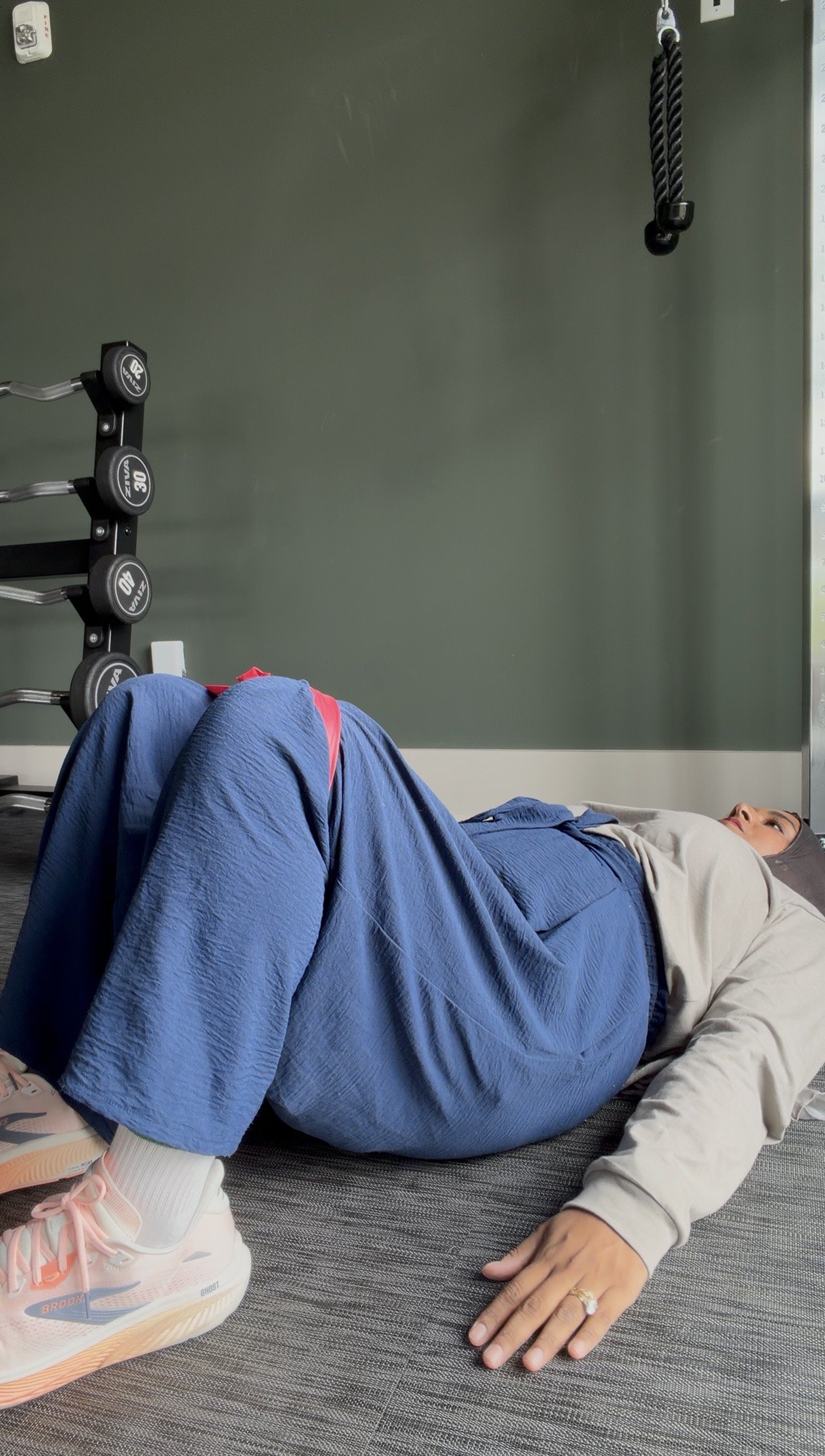 A woman lying on her side on a gray mat in a gym, resting with her eyes open, wearing a headscarf, gray sweatshirt, blue pants, pink and white sneakers, with weightlifting equipment and a rope hanging from the ceiling in the background.