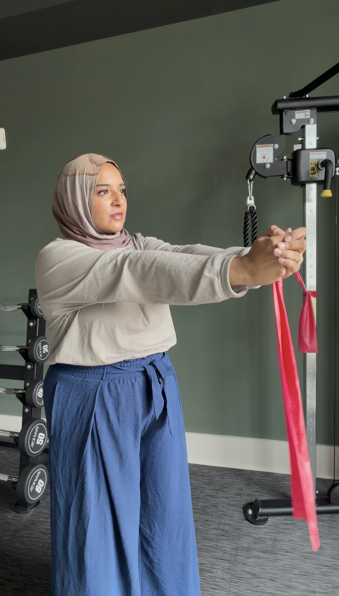 A woman in a beige hijab and blue pants working with resistance bands in a gym.