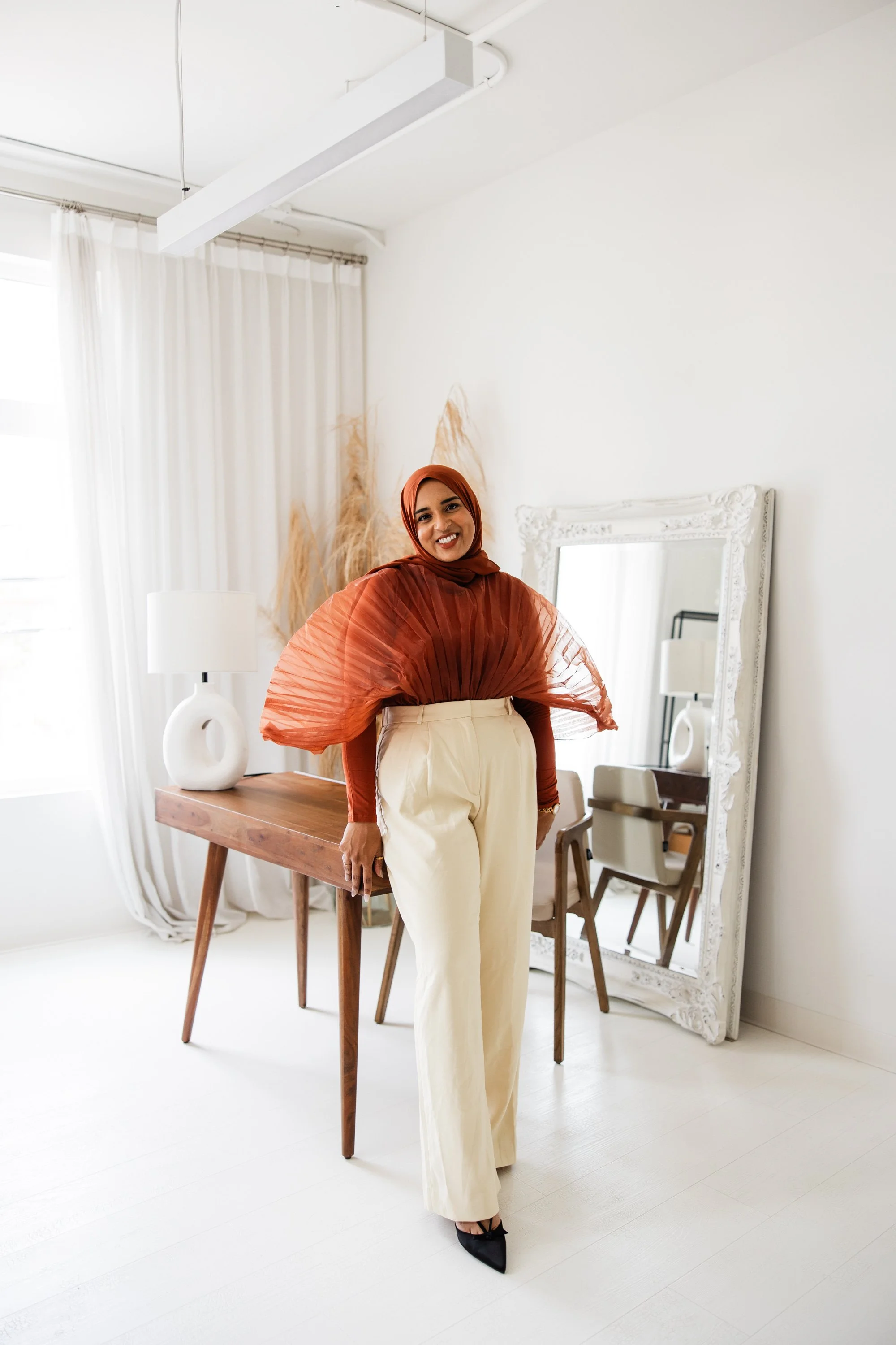 A woman smiling in a modern, minimalist room with white walls, beige curtains, a wooden console table, a white vase, a mirror, and a chair.