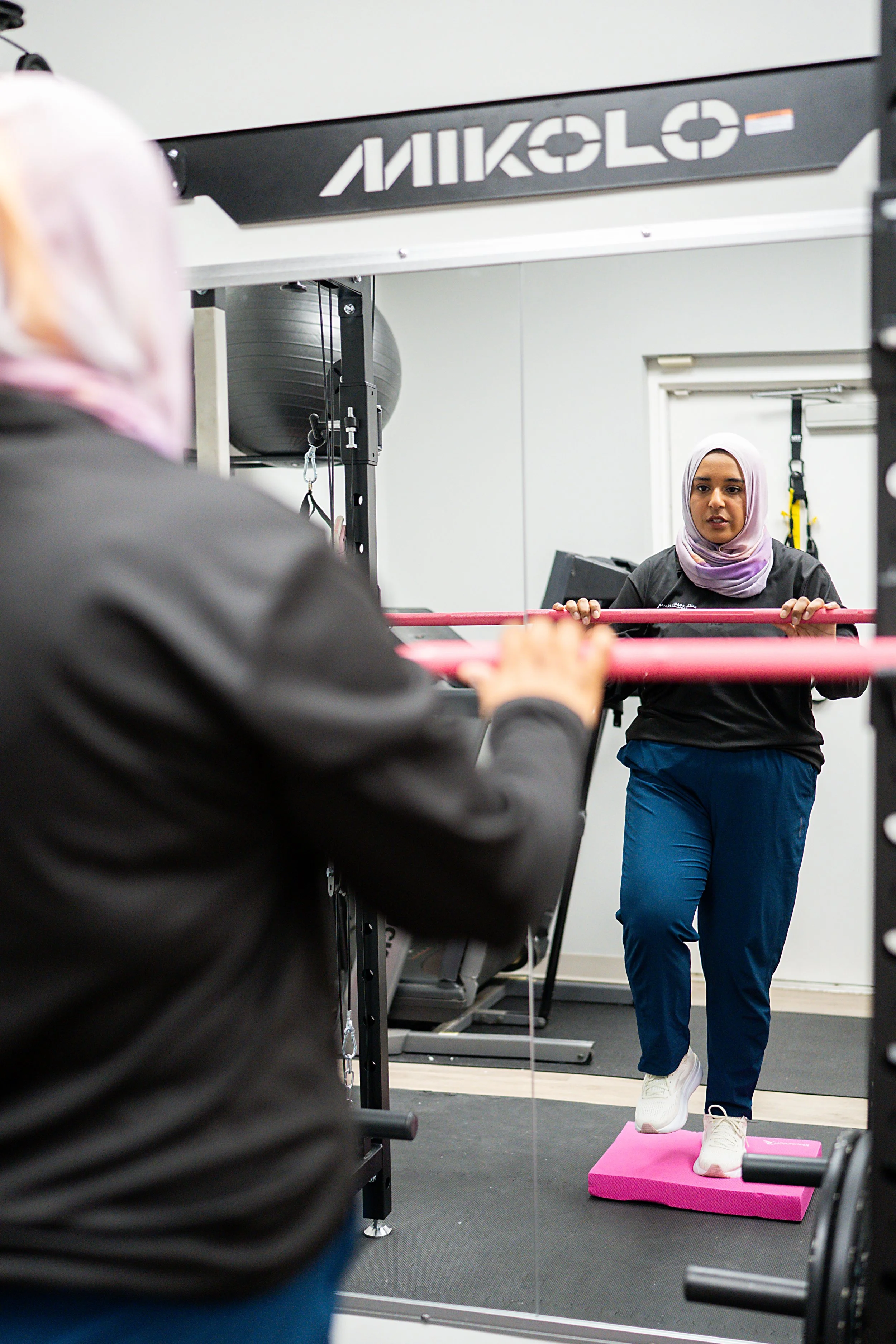 A woman in a hijab and athletic clothing performing an exercise on a pink step platform in a gym, with a mirror behind her reflecting the scene.