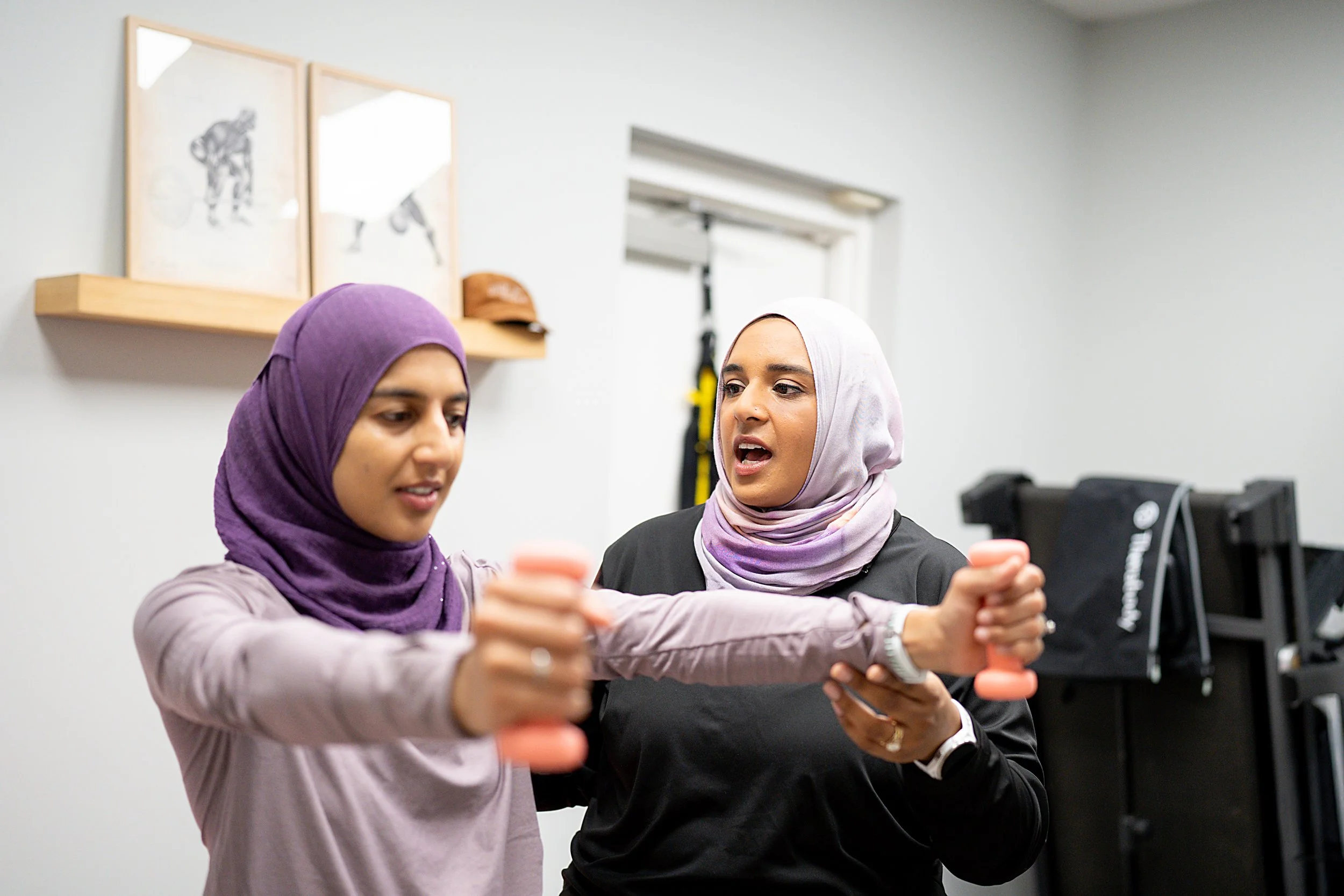 Two women wearing headscarves smiling and holding small dumbbells during a workout session in a gym.