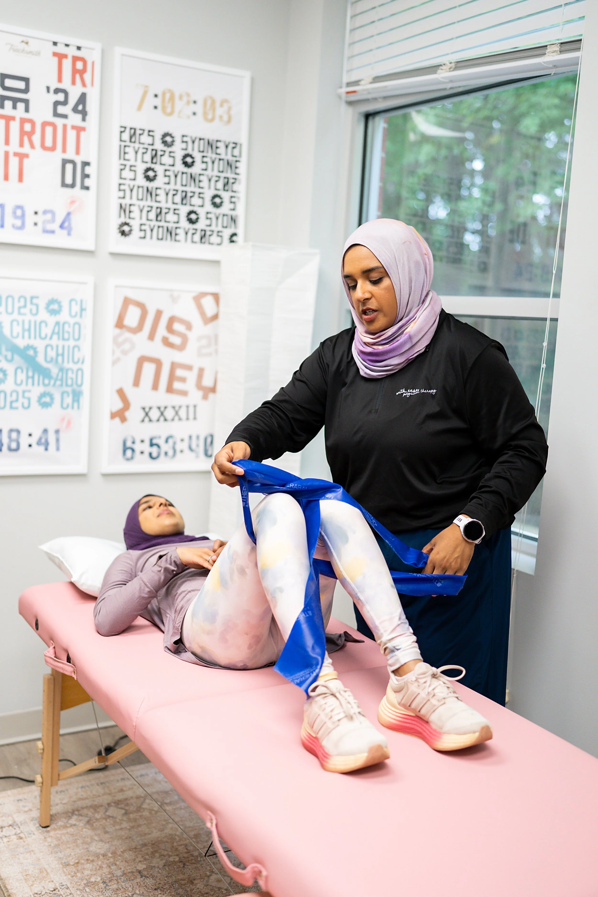 A woman with a purple hijab lying on a pink massage table, with her legs raised and a woman with a light purple hijab and dark shirt standing beside her, holding a blue sling. The room has posters of different race tracks and a large window showing trees outside.