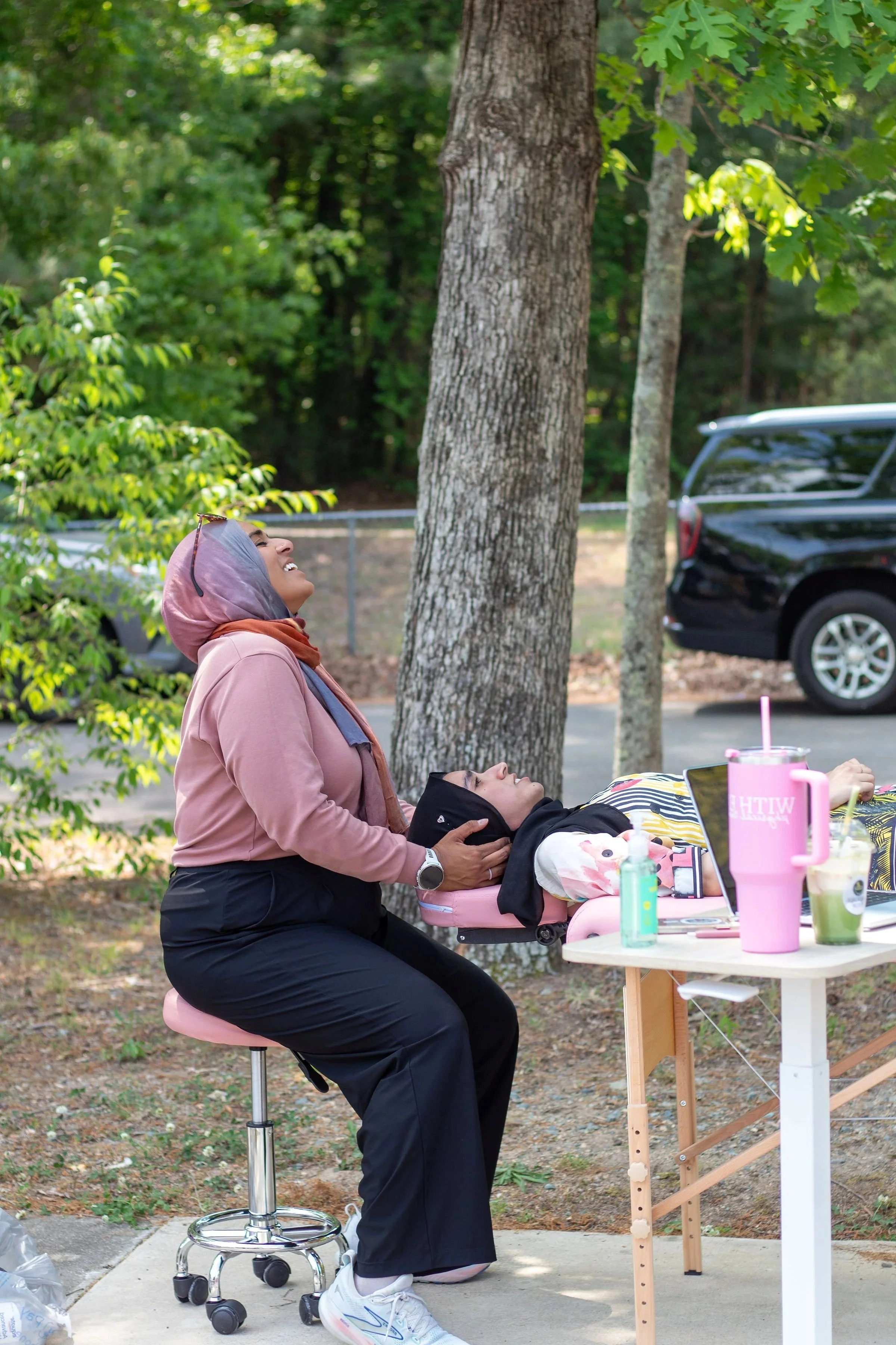 Woman in pink jacket and headscarf kneeling on the ground, holding a woman's head who is lying in a stroller, with a background of trees and parked cars.