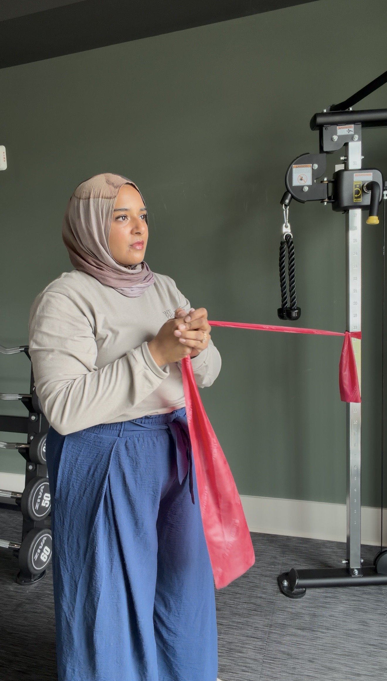 A woman wearing a beige long-sleeve top, blue pants, and a hijab, standing in a fitness studio with a weightlifting machine and weights behind her, holding a resistance band attached to the machine.