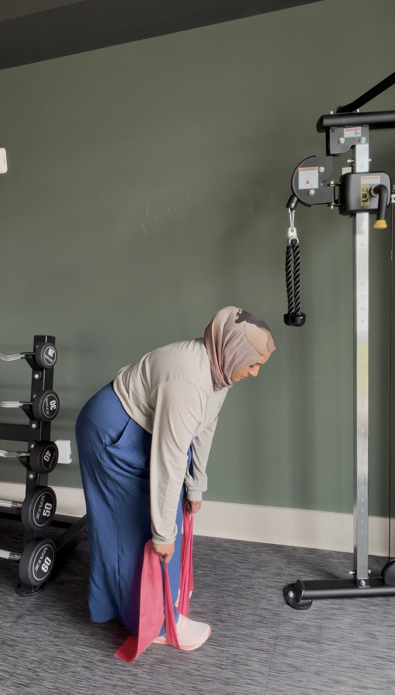 A woman in a beige hoodie and blue pants is participating in a fitness exercise at a gym, using resistance bands while bending forward.