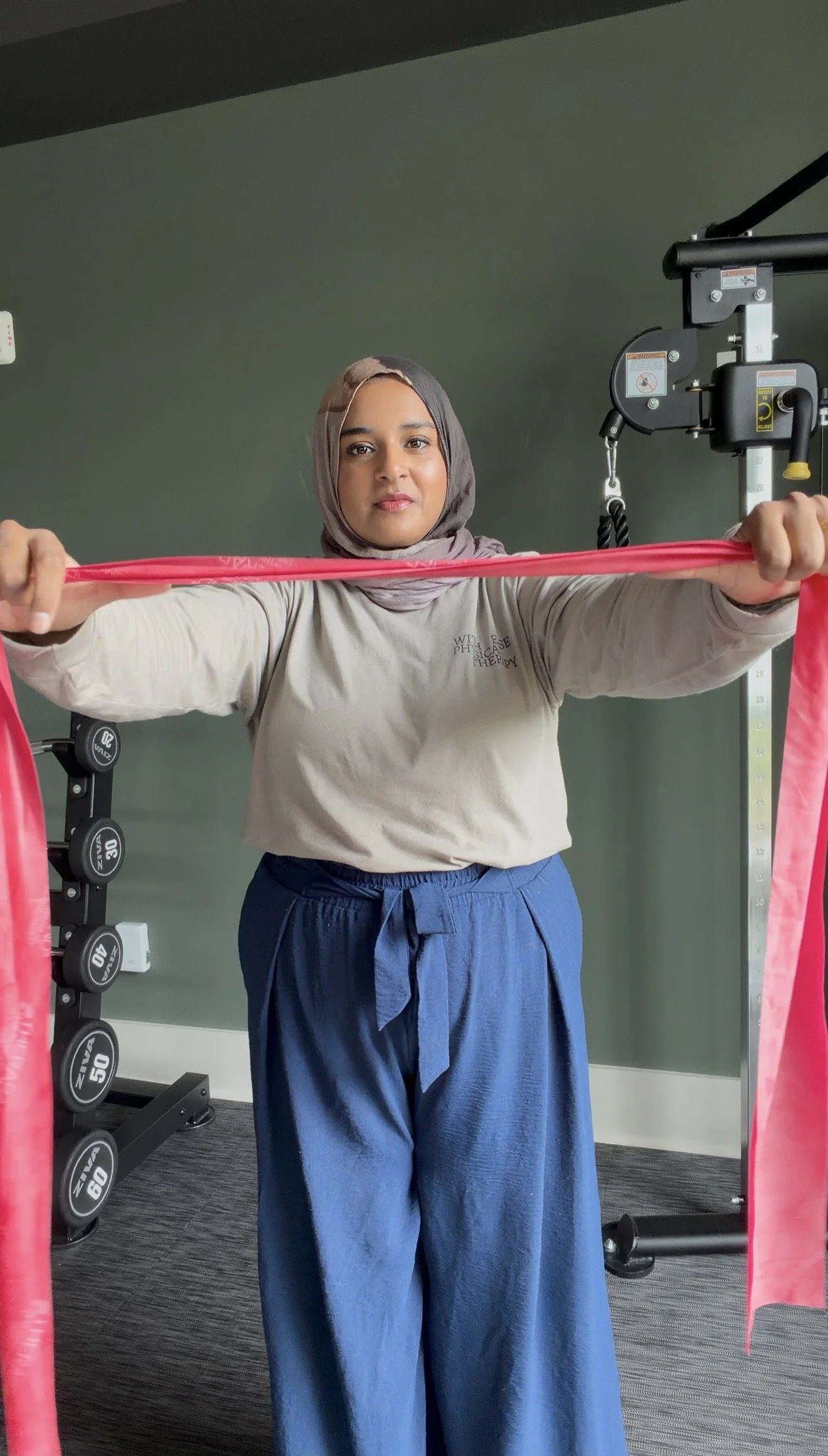 A woman wearing a hijab and beige long-sleeve shirt holding a pink resistance band in a gym with weightlifting equipment and green walls.