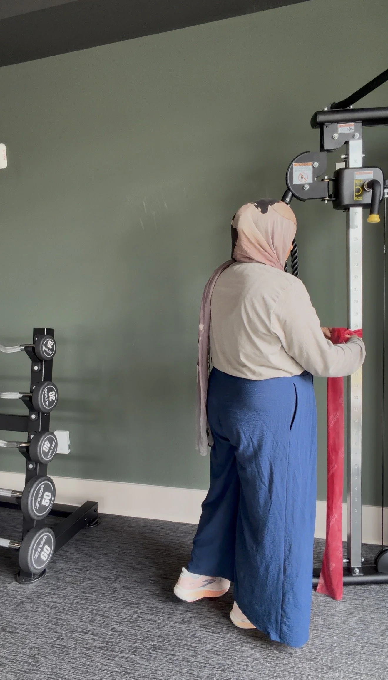 A woman with a light pink headscarf, beige top, and blue pants using a red resistance band attached to a white height measurement stand in a gym. There are weight plates on a rack to the left and a green wall in the background.