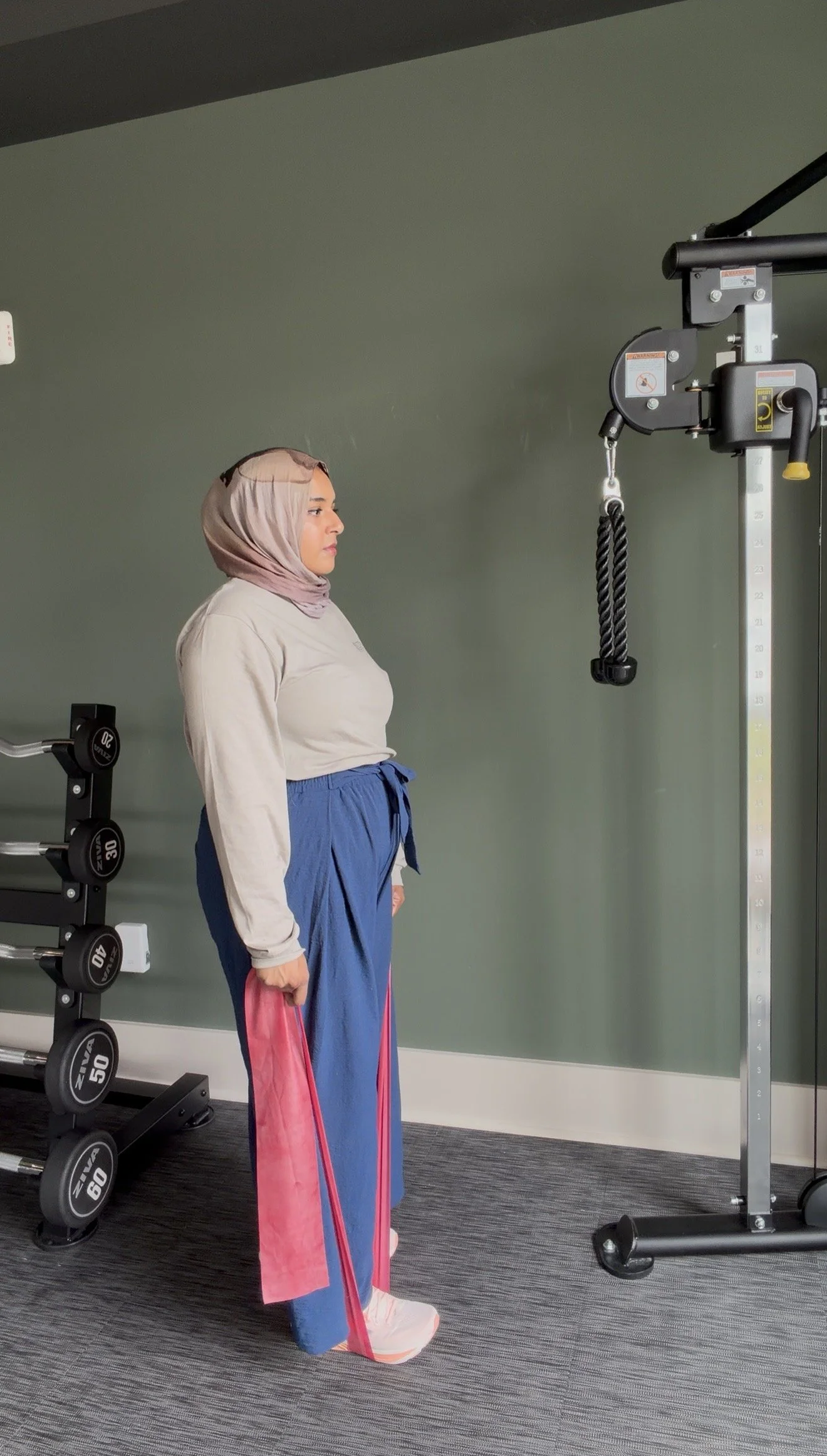 A woman wearing a beige hijab, gray long-sleeve shirt, blue pants, and pink gaiters holding a pink resistance band standing by gym equipment and a pulley machine in a gym with a dark green wall.