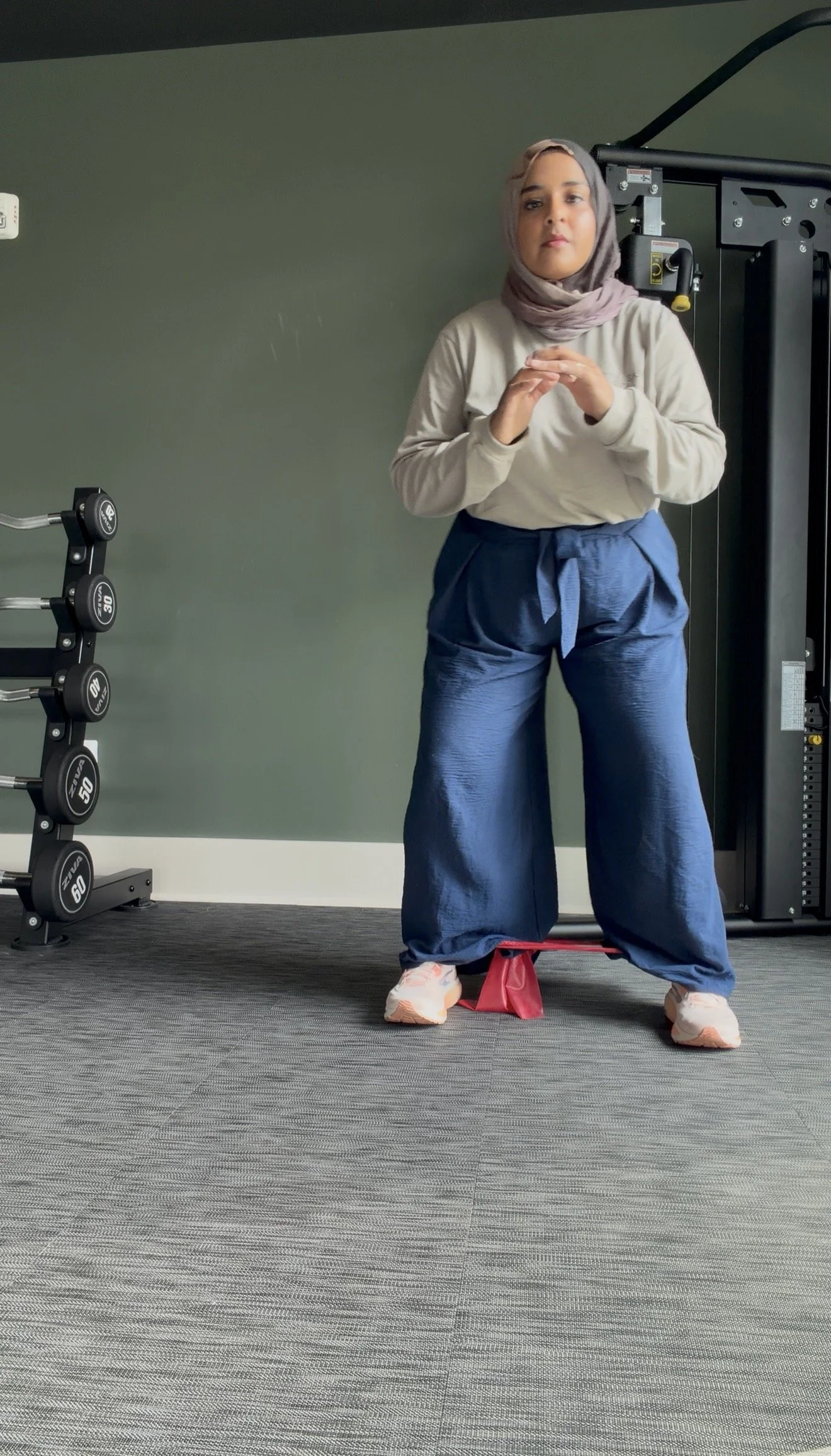 A woman wearing a hijab, beige sweatshirt, and blue wide-leg pants exercising on a balance board in a gym, with weights and exercise equipment in the background.