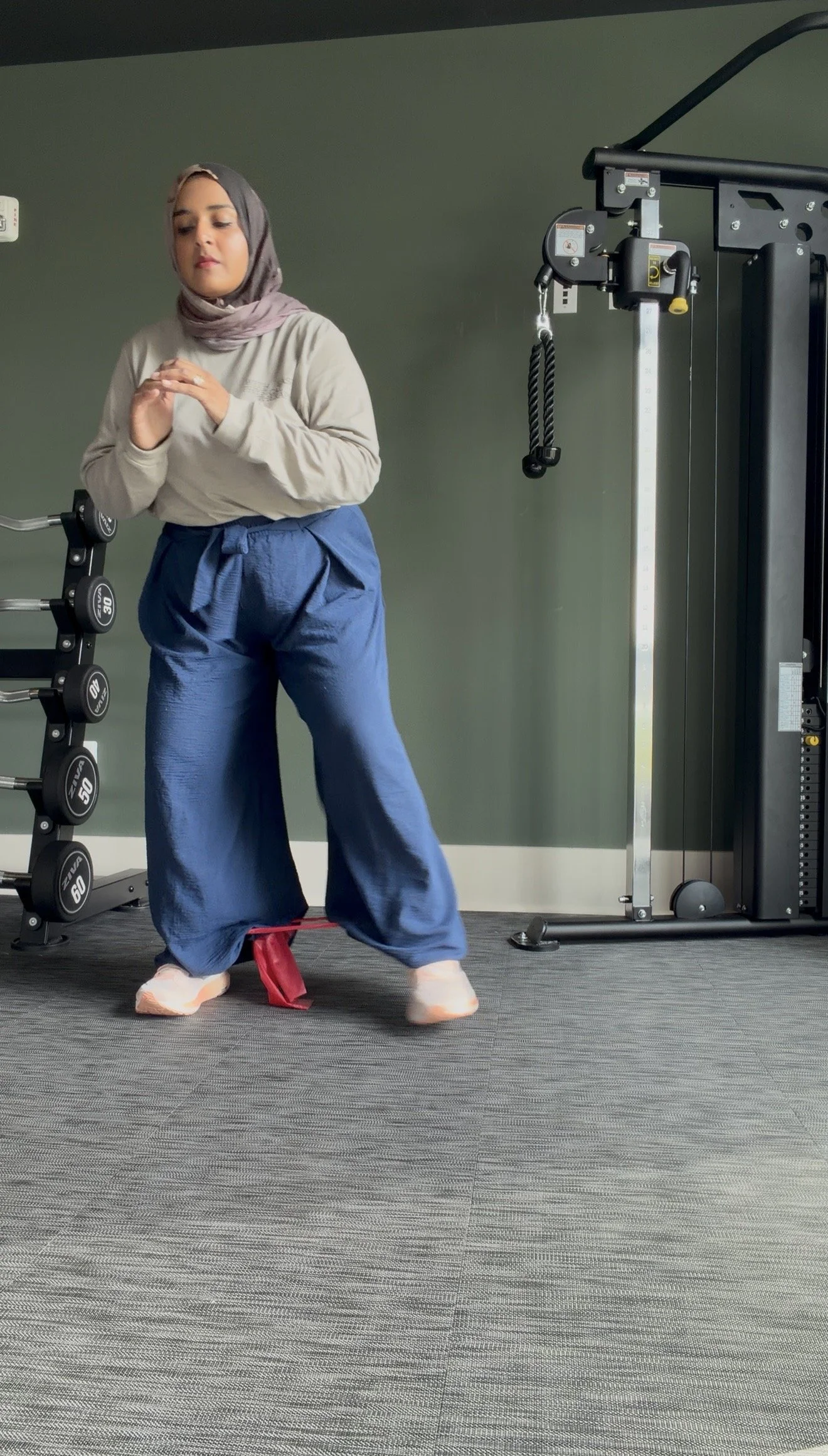 A woman in a gray hijab, beige long sleeve shirt, and wide-leg blue pants is exercising in a gym, balancing on a red balance pad while holding her hands together. There are workout equipment and a green wall in the background.