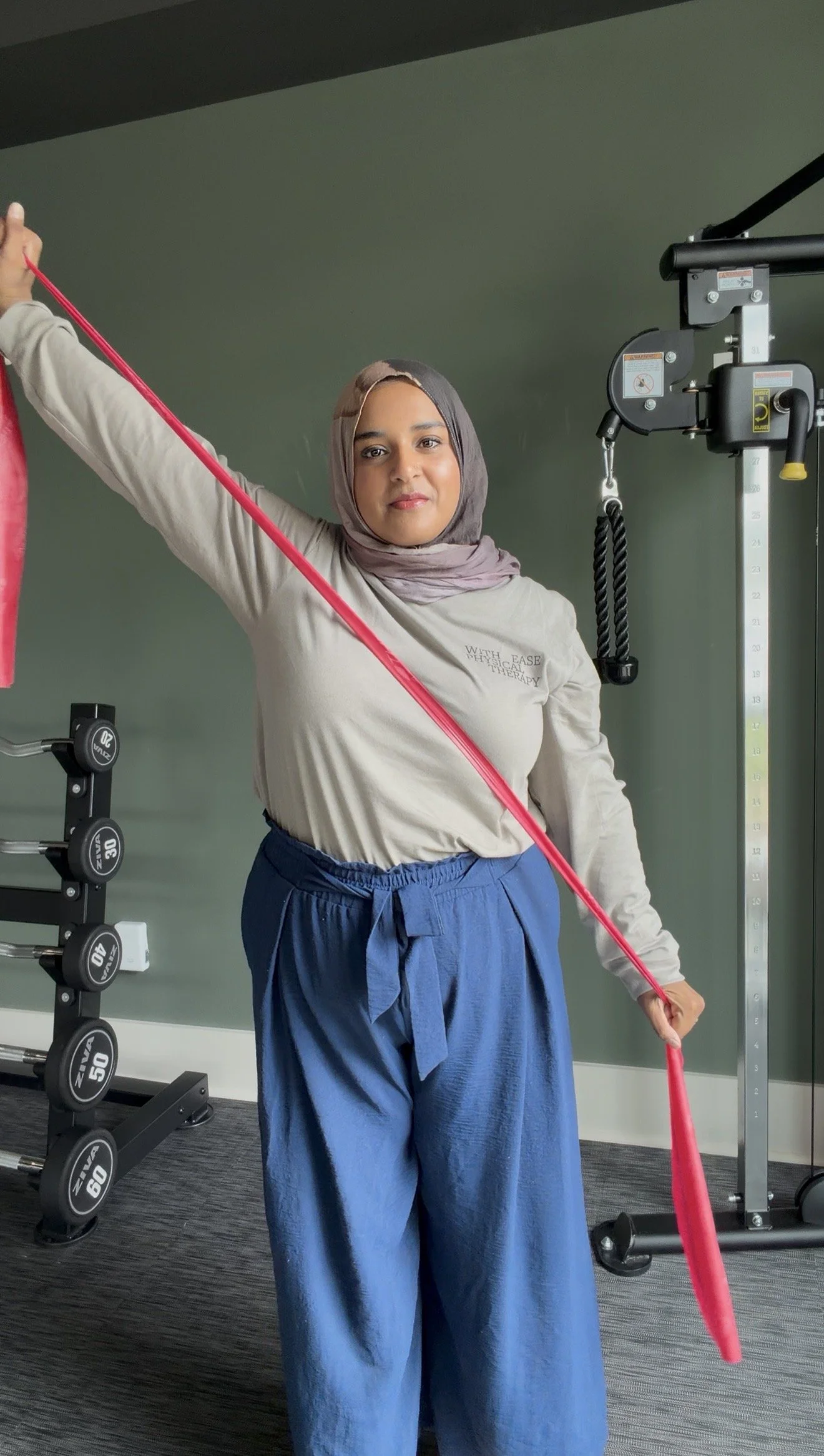 A woman wearing a hijab and loose blue pants exercising with a resistance band at the gym.