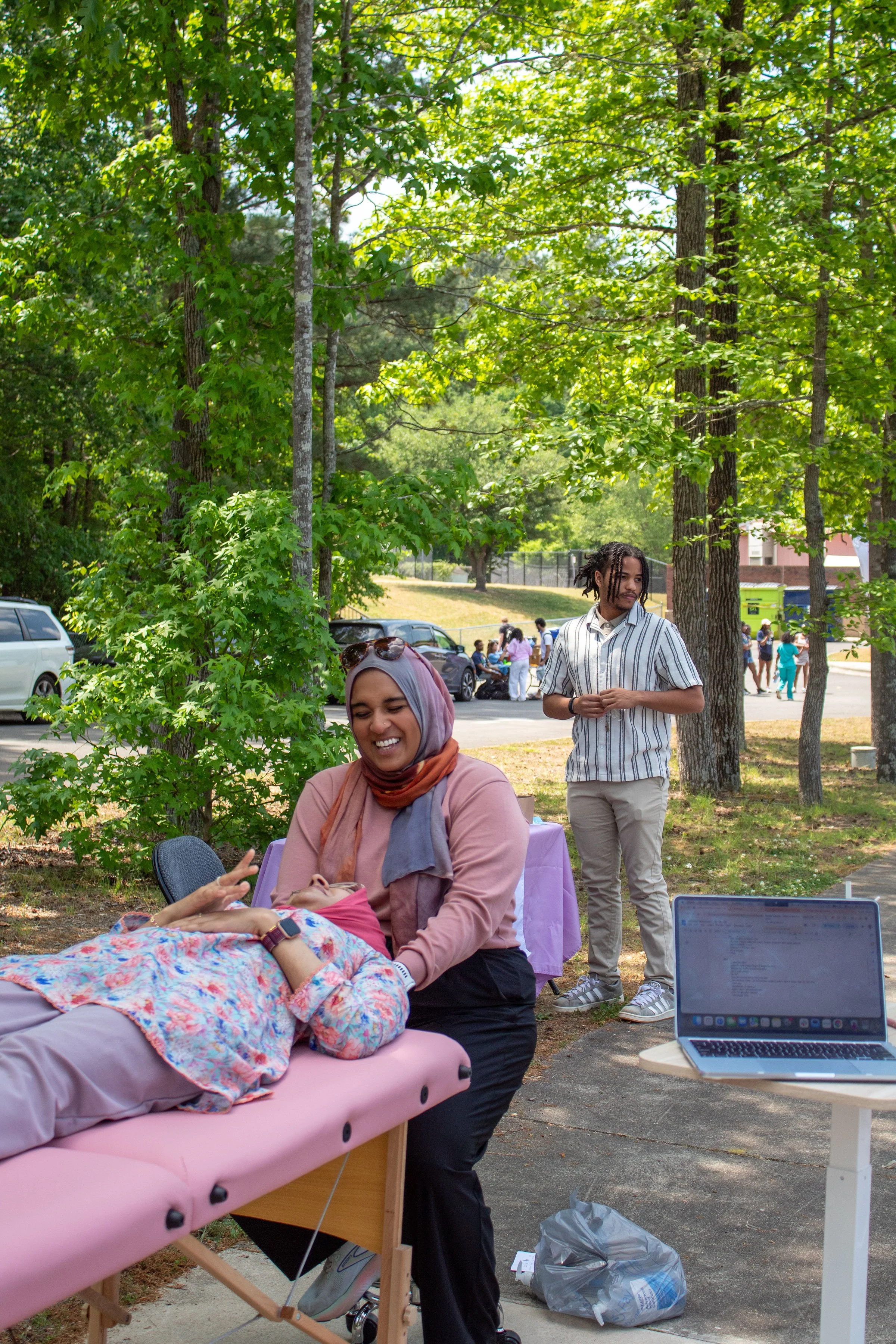 A woman in a hijab and a pink shirt is lying on a pink portable massage table with a colorful blanket covering her. She appears relaxed and is smiling, engaging with a person beside her who is not fully visible. Behind them, a healthcare worker with dreadlocks, a striped shirt, and beige pants is standing and observing. In the background, a man with dreadlocks, a striped shirt, and beige pants is standing with arms crossed, looking at the woman on the table. The scene takes place outdoors under trees, with cars parked in the background and a group of people gathered nearby. There is a laptop on a small white table to the right, and a plastic bag on the ground nearby.