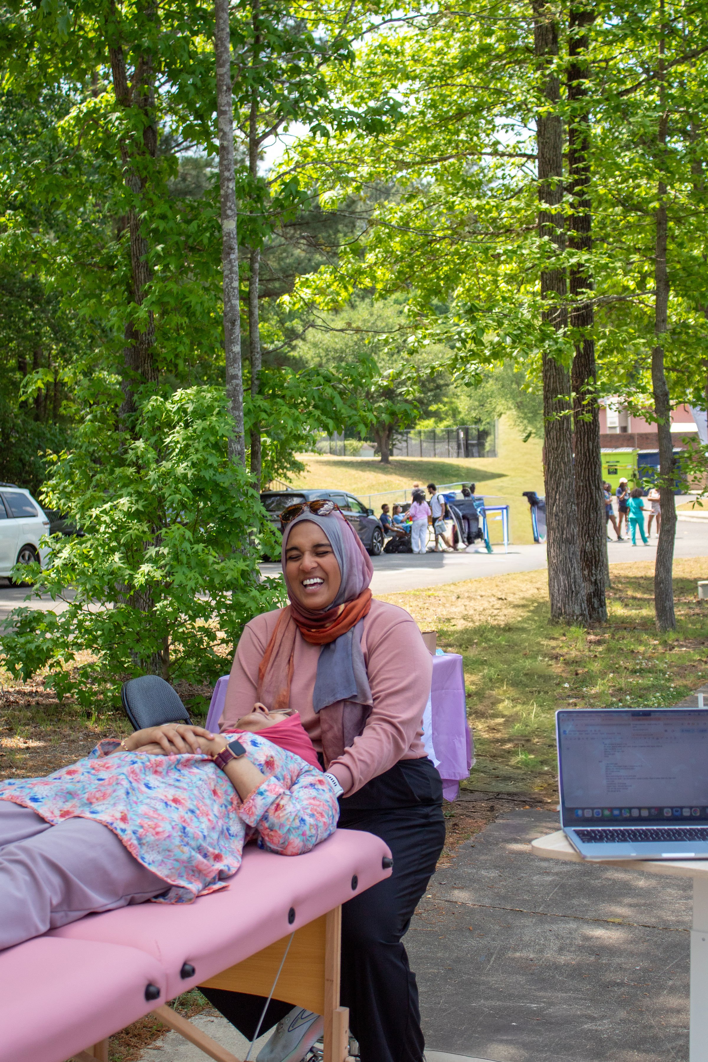 A woman in a hijab smiling and kneeling next to a person lying on a massage table outdoors. The person on the table is covered with a floral blanket. There is a laptop on a table nearby, and people in the background near a school bus and parked cars. Trees and a grassy hill are also visible.