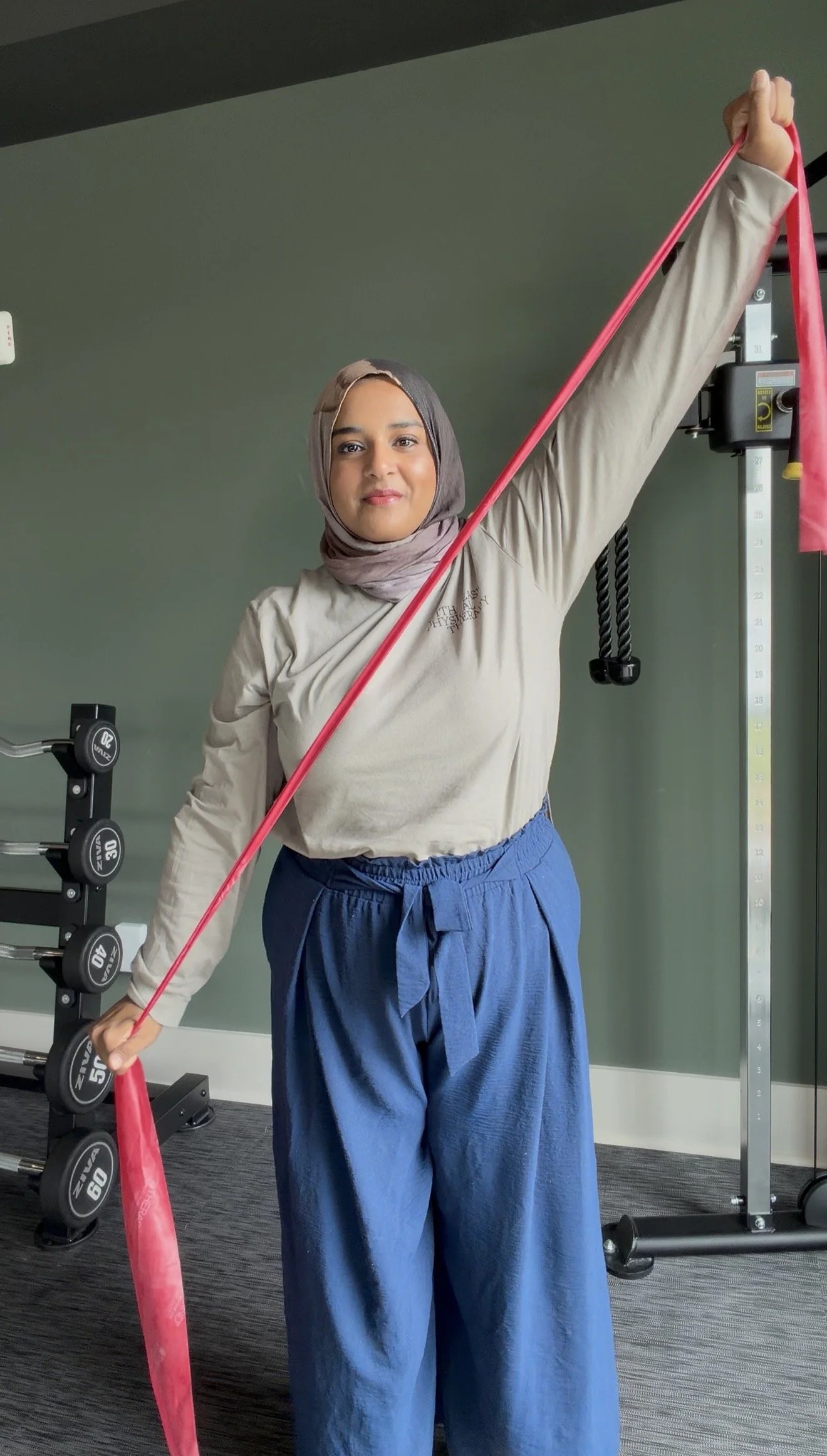 A woman wearing a beige long-sleeve shirt, blue high-waisted pants with a tied waistband, and a gray hijab standing in a gym. She is holding a pink resistance band with one arm extended overhead and the other arm down, with gym equipment visible in the background.
