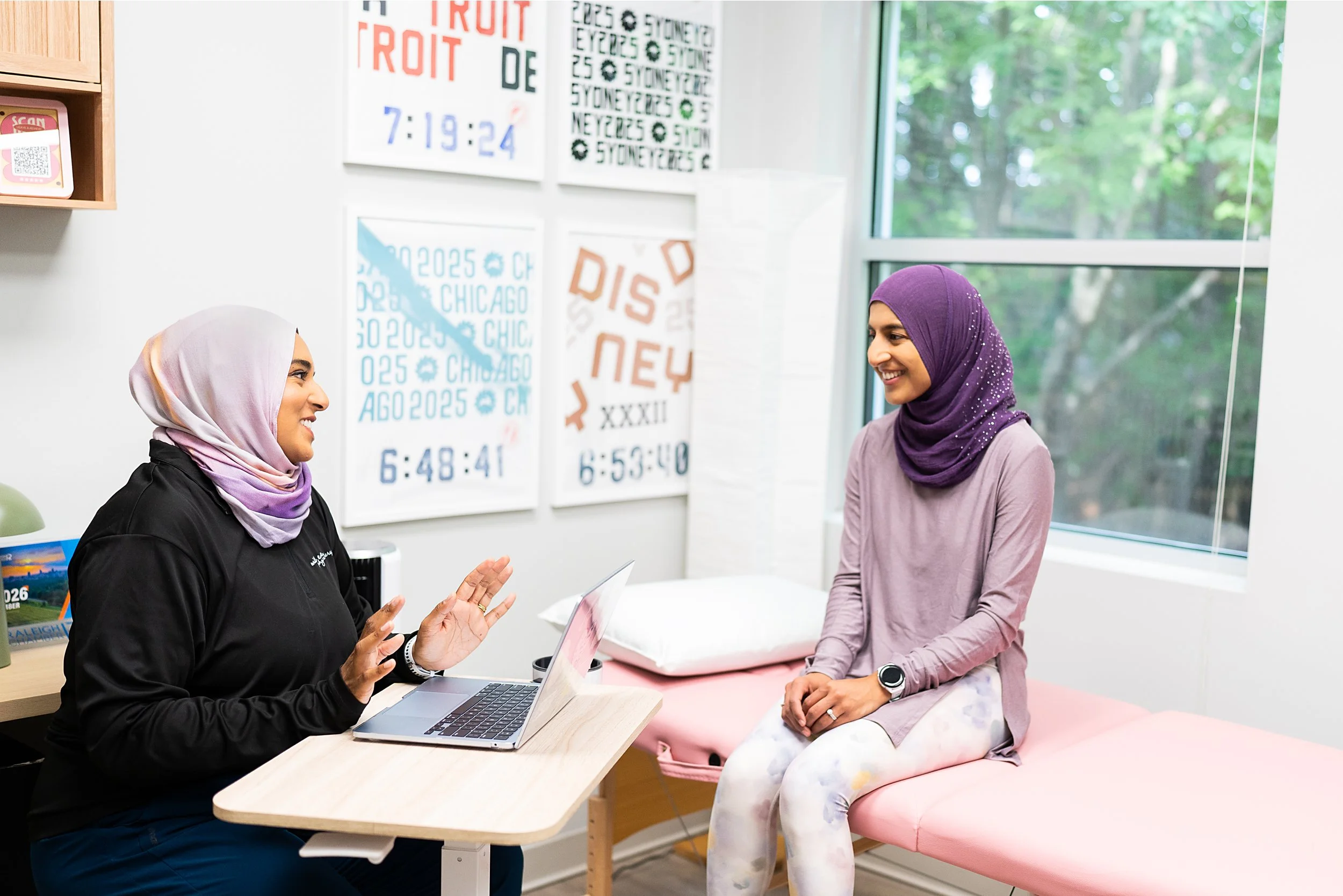 Two women wearing hijabs and smiling while having a conversation in a bright, modern office with large windows and colorful wall posters.