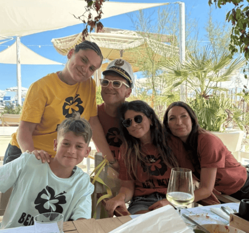 Gruppo di cinque persone sedute a un tavolo all'aperto, sorridenti, con due bambini e tre adulti, in un ambiente soleggiato con piante e decorazioni.